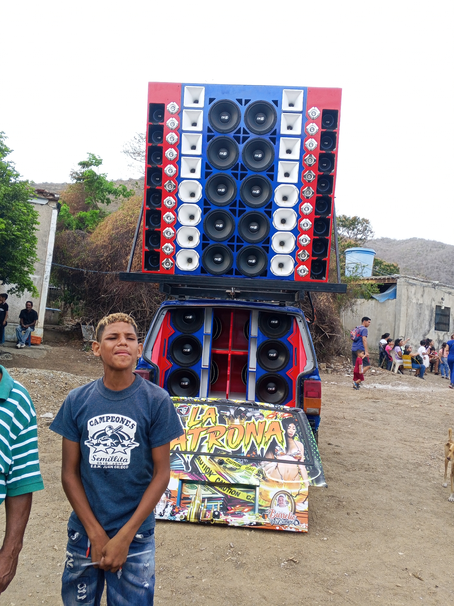 The image captures a scene in what appears to be Santa Ana, Venezuela, although the exact location within the city isn't specified. In the foreground stands a young boy wearing a blue t-shirt that reads "Campeones" with the word "Semillita" and "E.B.M. Juan Griego" underneath, paired with jeans. He stands in front of a blue SUV with an enormous, custom-built sound system mounted on top and inside the car. The sound system is adorned in red, white, and blue and features numerous speakers. The back of the SUV has an advertising panel featuring a woman in swimwear and the words "La Patrona", with smaller text including the phrase "Burrelio Por Siempre".
In the background, there's a group of people sitting and standing around, suggesting a gathering or event. The setting includes houses, trees, and a dusty or dirt ground, typical of a less developed urban area. A dog is also visible on the right side of the frame. Overall, the scene captures a cultural moment involving a large sound system often seen in Venezuelan street celebrations.