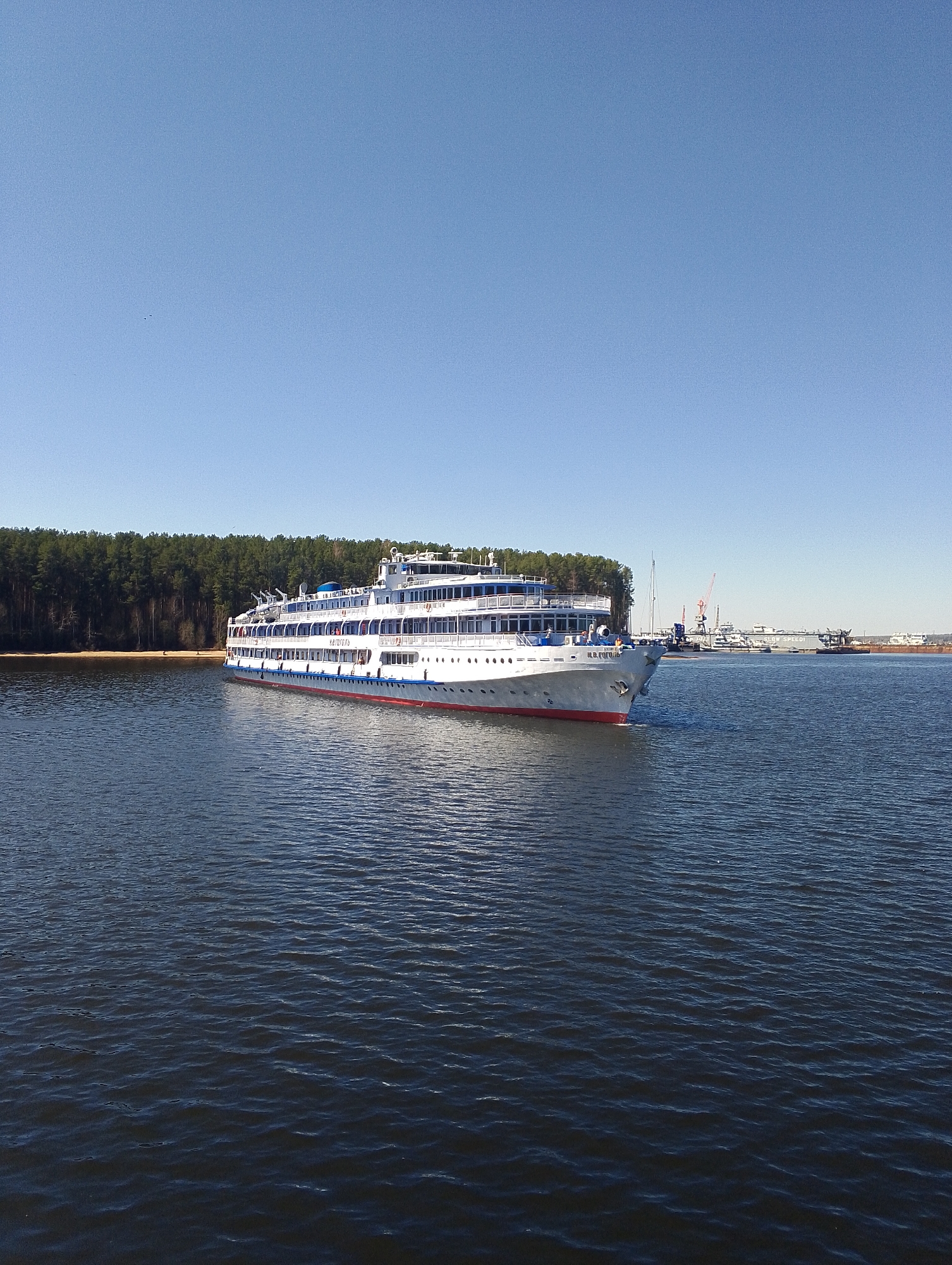 A large, white passenger ship with a red stripe at the waterline is sailing on the Kama River in the city of Chaikovsky, Russia. The ship features multiple decks and several windows on each level. The bow of the ship is facing towards the right side of the frame.
Behind the ship, on the left side of the frame, is a dense forest extending along the shoreline. The background on the right shows a port area with cranes, docked ships, and other harbor structures. The sky above is clear and blue. The water is dark and rippled.