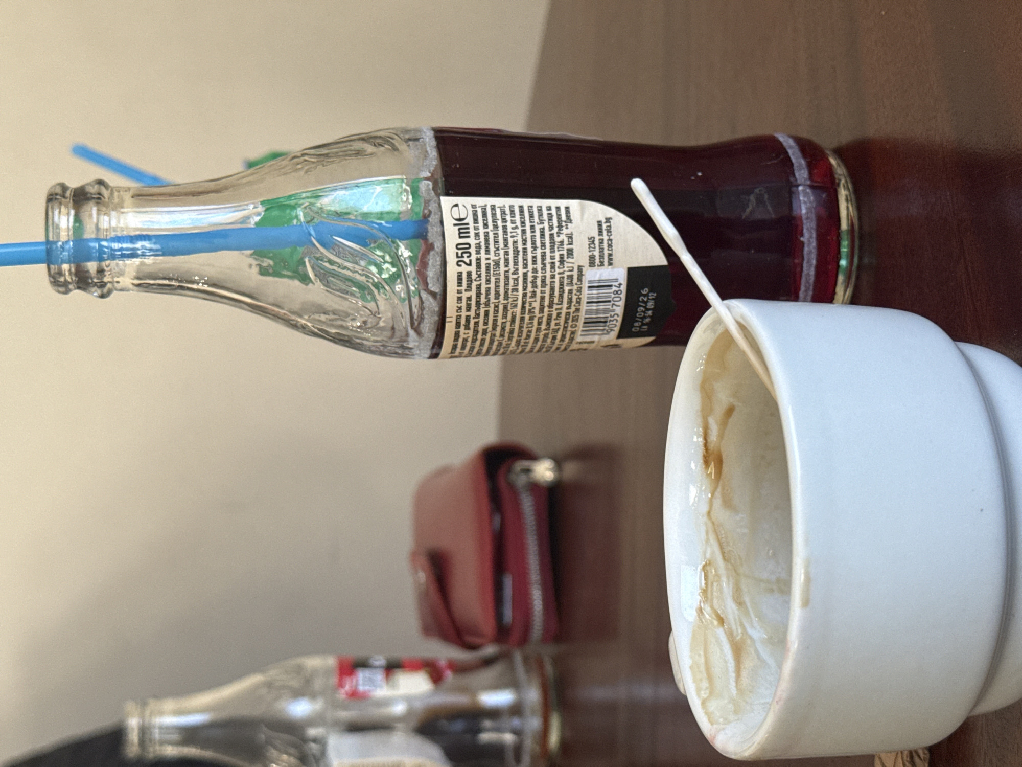 The image shows a table with various objects. There's a half-empty Coca-Cola bottle with a blue straw sticking out of it. Next to it is an empty, white coffee mug with coffee stains inside. A red wallet is also present, slightly out of focus in the background. A second, empty bottle is barely visible in the bottom left corner. The setting appears to be indoors, possibly in a café or restaurant. The Coca-Cola bottle label indicates it is a Bulgarian product, with the contact information being in Sofia, Bulgaria, but does not indicate the location of Пампорово. The image lacks any information that connects it to the city of Пампорово.