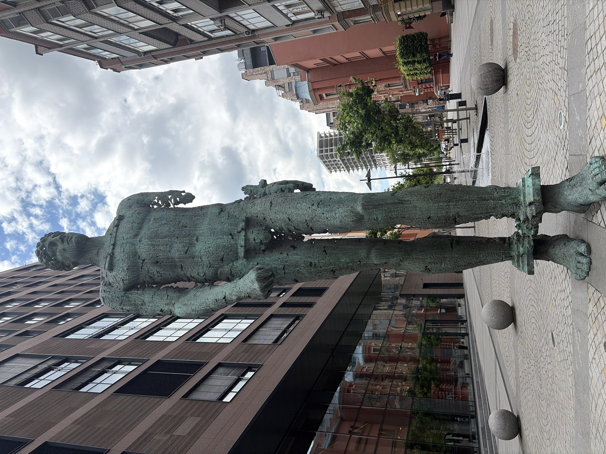 The image shows a full-length bronze sculpture of a male figure standing in a city square in Dnipro, Ukraine. The sculpture is approximately twice the height of a human and is weathered with a greenish patina. It is missing its right arm from the shoulder down and its left arm from the elbow down, and its genitals are exposed. The sculpture stands on a pedestal, and its feet are bound by metal bands.
The square is paved with cobblestones. In the foreground, to the left and right of the sculpture, are two large, spherical stone bollards. Behind the sculpture, a modern building with a glass facade and rows of windows is visible. Reflections of red brick buildings can be seen in the glass.
Further in the background, a street is lined with trees and buildings. Several people are walking on the sidewalk, and a fountain is active, with water spraying. Cars are parked along the street, and a construction site with an unfinished building is visible in the distance. The sky is cloudy with patches of blue. The overall scene depicts an outdoor urban environment.
