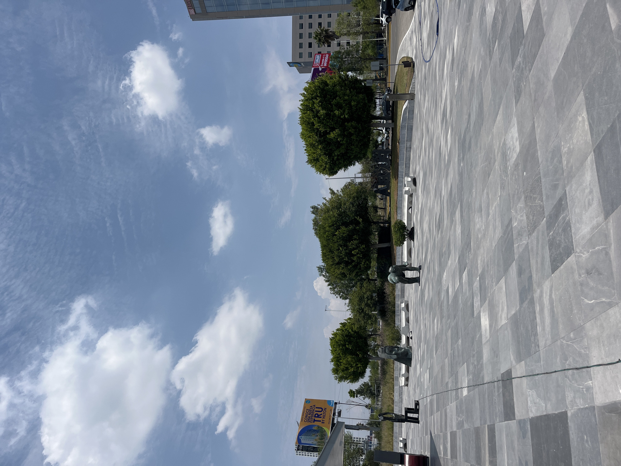 The photograph depicts a public space in San Bernardino Tlaxcalancingo, Mexico. The foreground is dominated by a wide, tiled plaza with a geometric pattern. Three sculptural figures are positioned along the plaza's edge near trees and grass. People are walking around these statues. Beyond the plaza, there are additional trees, some signage including a billboard for Tru by Hilton, and buildings including a multistory hotel. The sky is blue with scattered white clouds.