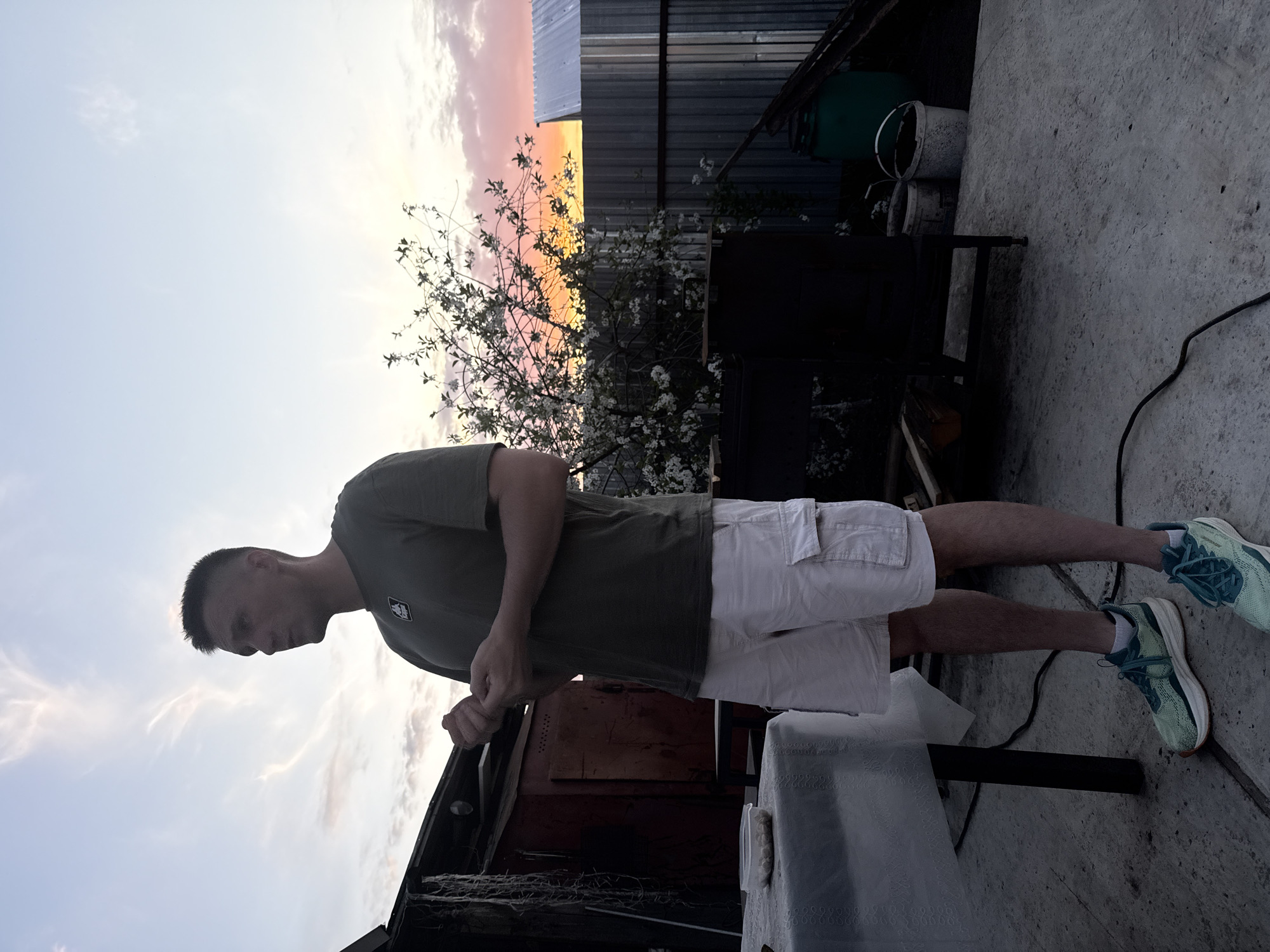 A full-body shot of a man standing in an outdoor setting, likely a backyard or similar area. The man is wearing a dark green t-shirt, light-colored cargo shorts, and light blue athletic shoes with dark blue laces. He is looking down, with his hands positioned in front of him, possibly resting on a table.
Behind the man, there is a structure resembling a shed or small building with a rusted metal exterior. There is a table covered with a white tablecloth to the right of the shed. Above and behind the shed, there is a flowering tree or shrub in bloom, set against a vivid sky with colors ranging from white to orange to purple, suggesting it is either sunrise or sunset.
In the background, there is a metal fence, and above that, a metal roof. There are also a couple of buckets and other assorted items. The ground is concrete.
The location is speculated to be in the city of Chaikovsky, Russia.