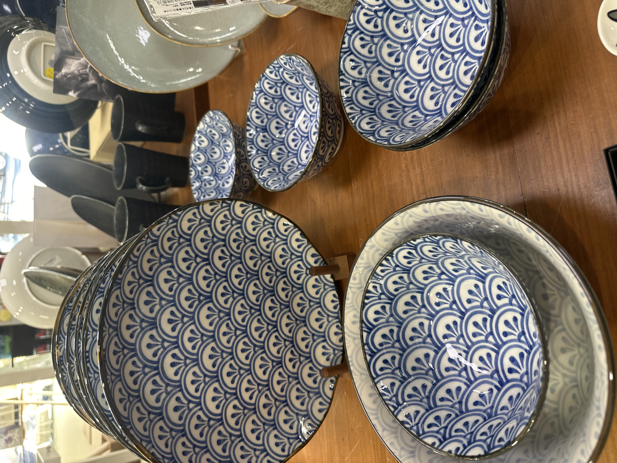 This is a medium-shot, indoor, eye-level photograph of a display of blue and white patterned ceramic tableware on a wooden surface. The most prominent items are a stack of large plates with a repeating fan or scale-like pattern, and several smaller bowls with the same pattern. In the background, there are other ceramic items, including mugs and larger plates, some of which are out of focus. The lighting is neutral, and the overall impression is of a retail display. There are no people visible in the image. The image does not contain any information that would allow its location in Bellingen, Australia to be determined.