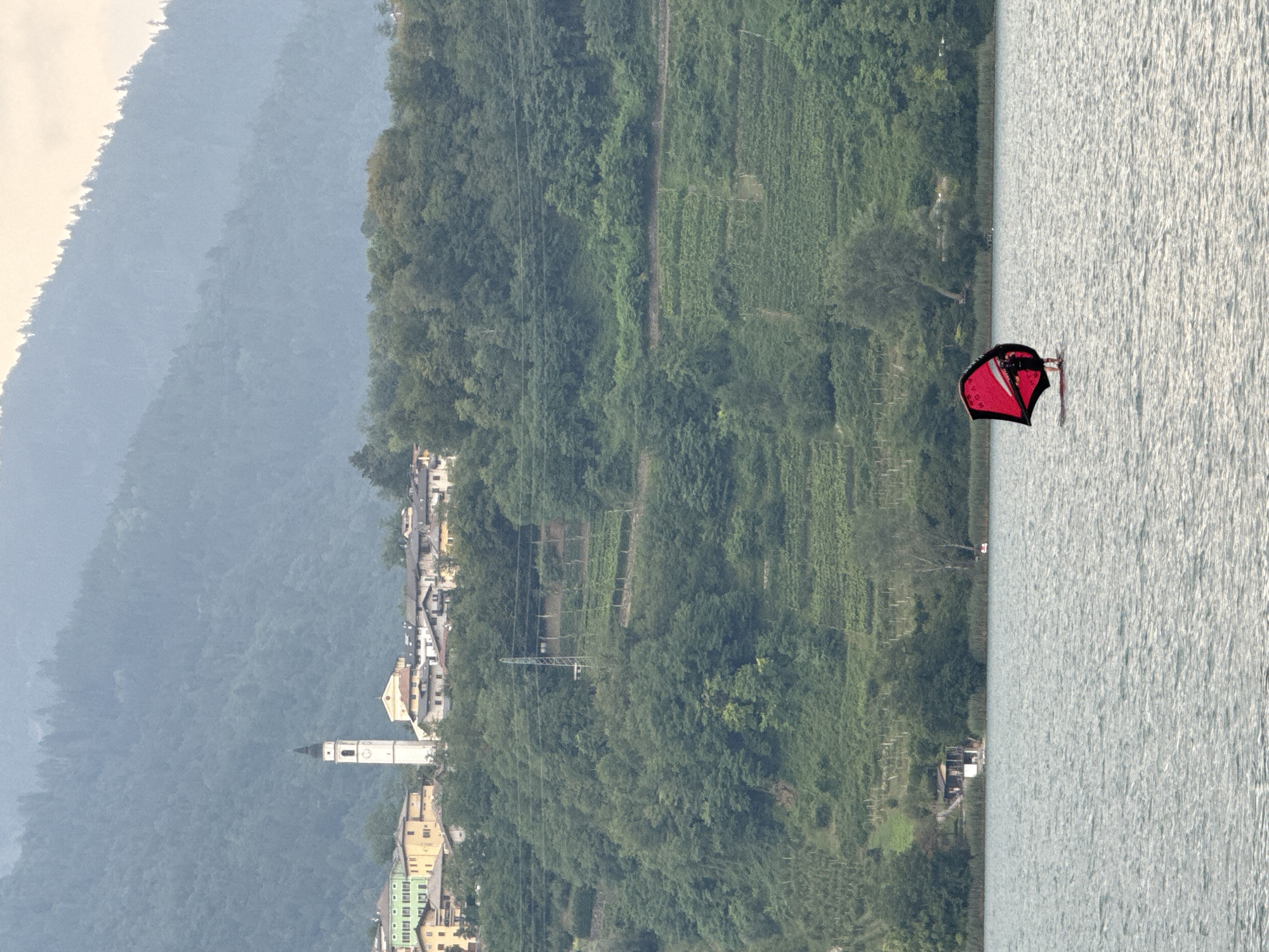 The image captures a serene view of Calceranica al Lago, Italy, on a cloudy day. In the foreground, a person is engaged in wing foiling on the shimmering lake. The foilboarder is equipped with a red wing, a type of wind-powered hydrofoil. The middle ground reveals a lush green hillside dotted with trees and terraced vineyards. A small, rustic building with a dark roof is situated near the water's edge, nestled amongst the reeds and trees. Further up the hillside, a cluster of houses forms a small village, characterized by various building colors including yellow, cream, and green. Dominating the village skyline is a tall, white church tower with a dark spire and a clock face. In the background, misty, tree-covered mountains rise, their peaks fading into the overcast sky. The overall scene is tranquil, highlighting the natural beauty and the presence of human activity within the landscape.