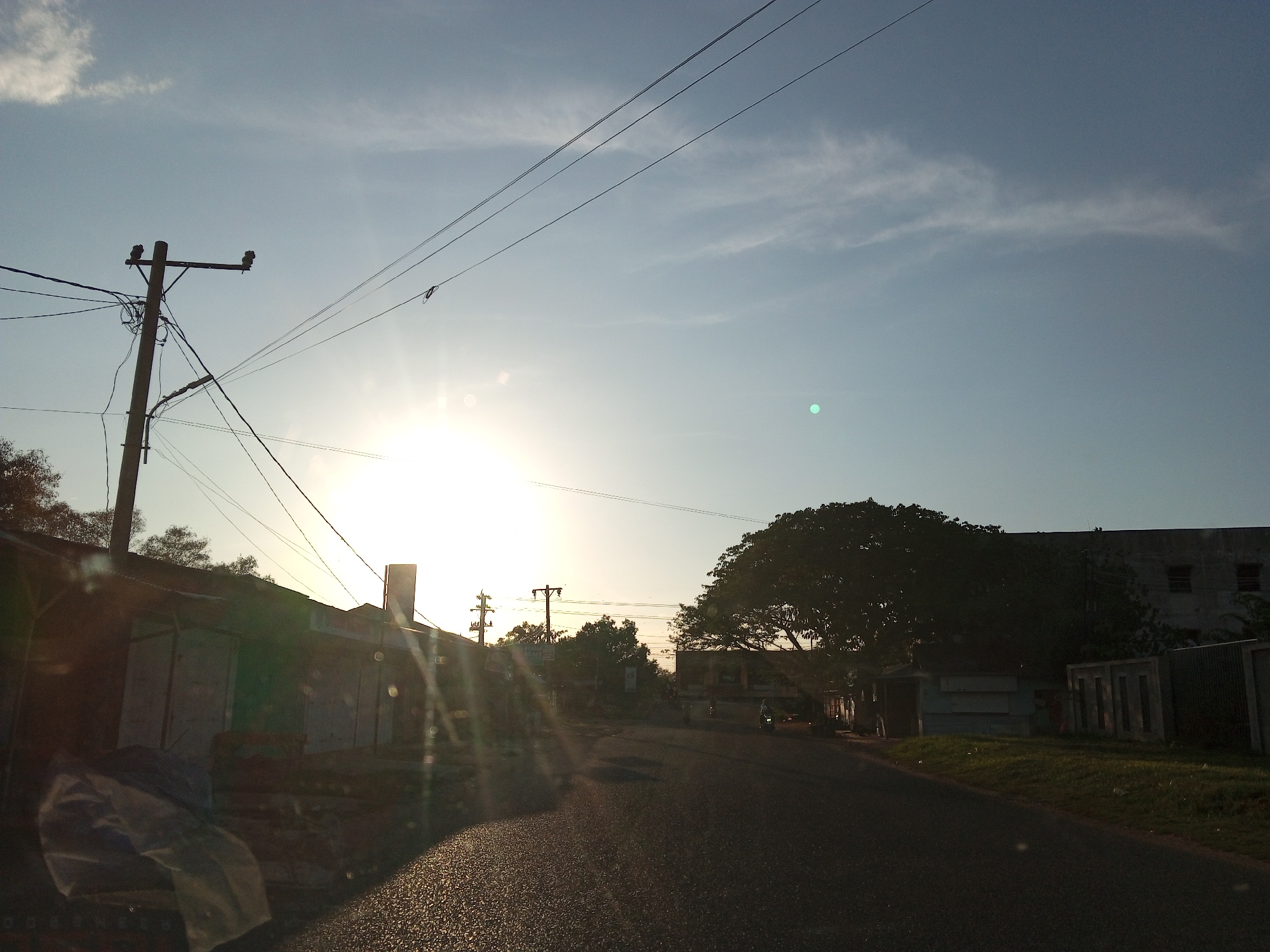 The image depicts a street scene likely in Lhokseumawe, Indonesia, based on the tropical setting. The time appears to be sunrise or sunset, with the sun as a bright spot on the horizon, partially obscured by buildings and trees. A paved road stretches into the distance, reflecting the light. Power lines crisscross above the road. Utility poles stand on either side of the street. Buildings line the left side of the road, some appearing to be small shops or houses, while a larger building is on the right, partly obscured by a large tree. No people are clearly visible, though there is a shape that could be a person, or a vehicle on the horizon.