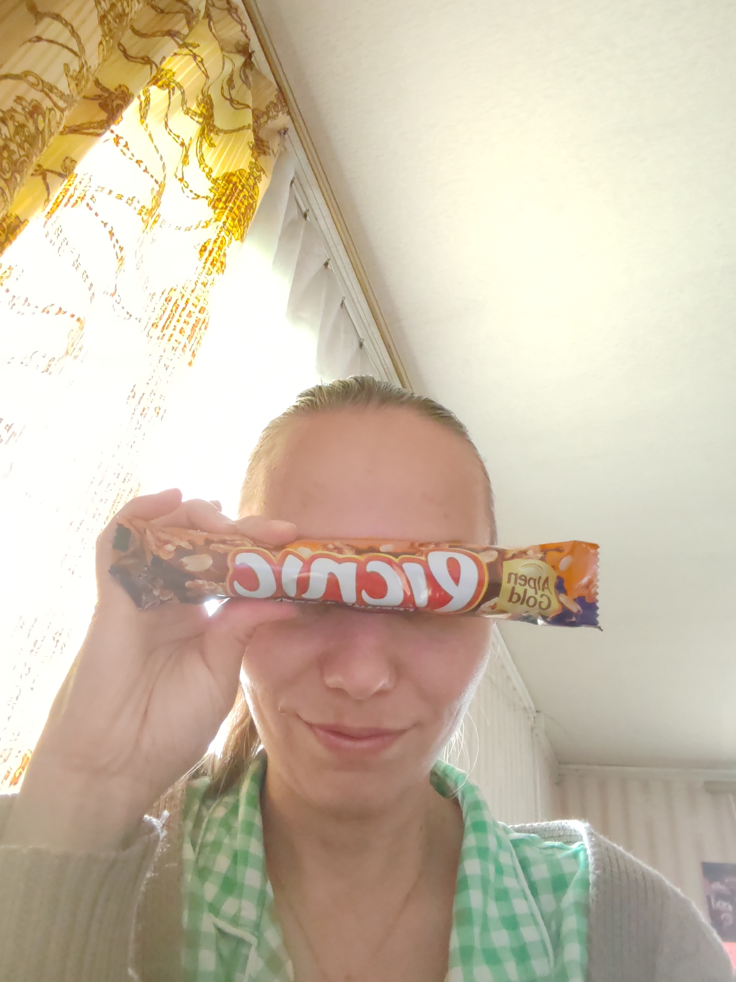 A woman is holding a chocolate bar over her eyes. The brand of the chocolate bar is "Alpen Gold". She is wearing a green and white checkered shirt and a beige sweater. In the background, there is a yellow curtain and a white ceiling. The woman is smiling slightly. The location is in Чухлэм, Russia.
