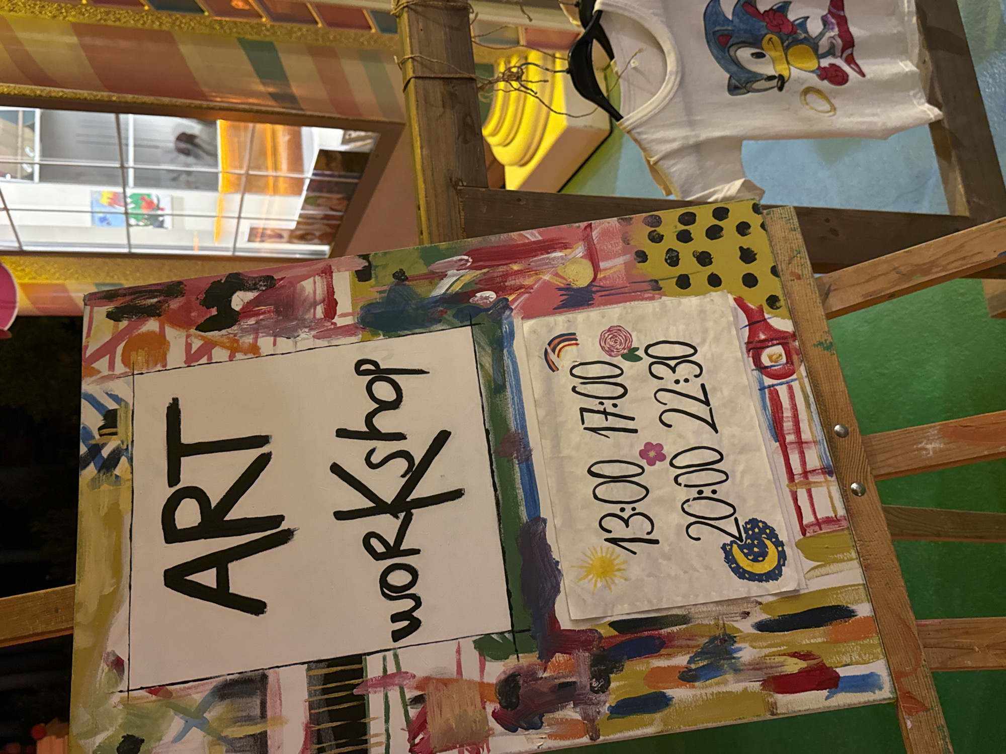 A sign with "ART WORKSHOP" painted in black letters on a white background sits on a wooden easel. Below the text, a smaller piece of paper displays times: "13:00 17:00" and "20:00 22:30", adorned with drawings of a sun, a flower, a rainbow, and a moon. The easel and sign are decorated with colorful abstract paint splatters. To the right of the easel, a white t-shirt featuring an image of Sonic the Hedgehog is displayed on a hanger. In the background, a window shows an indoor scene with shelves displaying various items. The overall setting appears to be an outdoor market or shop in Bodrum, Turkey, indicated by the presence of a child's t-shirt featuring a popular character, suggestive of a tourist-oriented location. The green surface below the easel could be artificial turf or carpet.