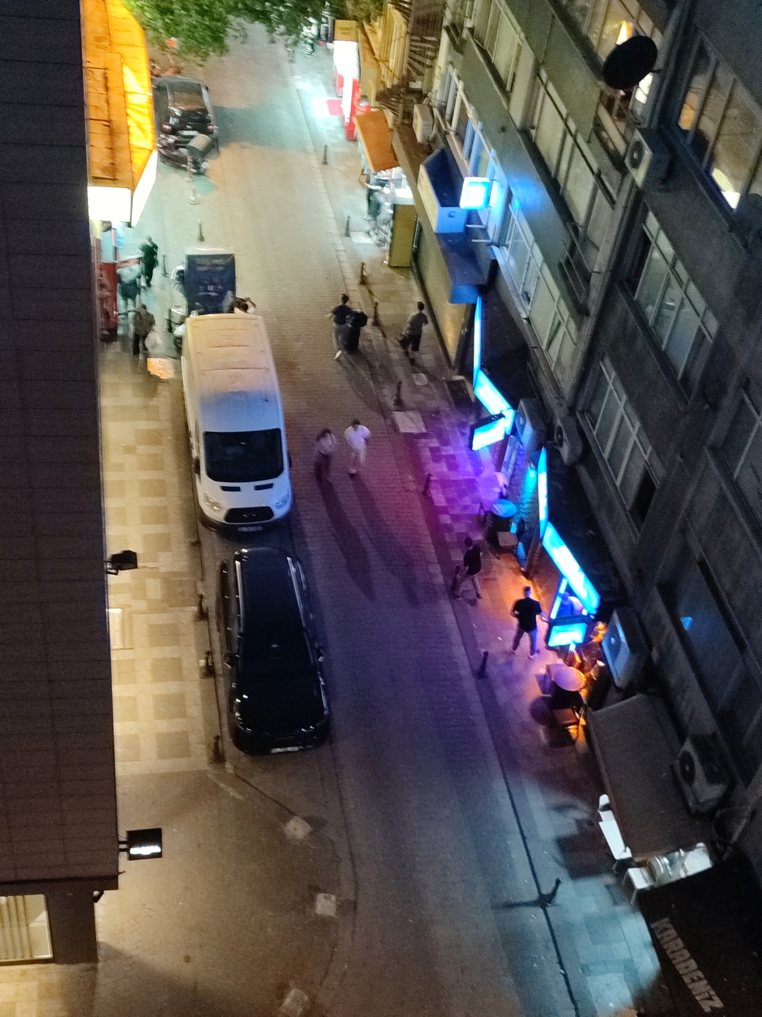 This is an overhead, night-time view of a street scene in Kadıköy, Turkey. A narrow street runs diagonally from the top left to the bottom right of the frame. On the left side of the street, a tall building with a dark, corrugated facade is visible. Adjacent to this, there is a section of a building with an illuminated yellow awning. The street itself is paved with cobblestones in some areas and asphalt in others.
Several vehicles are parked along the street. A white van is positioned in the middle of the street, and a black sedan is parked behind it. Further up the street, closer to the top left, a dark-colored car appears to be parked at an angle.
There are several people visible on the street and sidewalks. On the left side of the street, near the white van, a few individuals are standing and walking. On the right side of the street, adjacent to a multi-story building with numerous windows, there are several establishments with bright, blue neon lighting. People are seen standing in front of these establishments and walking on the sidewalk. The lighting from these establishments casts purple and orange glows on the street and sidewalk. In the bottom right corner of the image, a black object with white lettering that reads "KARADENİZ" is visible.
The environment suggests an urban setting with commercial activity, possibly shops or restaurants, illuminated by neon signs. The presence of streetlights and building lights creates a dynamic interplay of light and shadow.