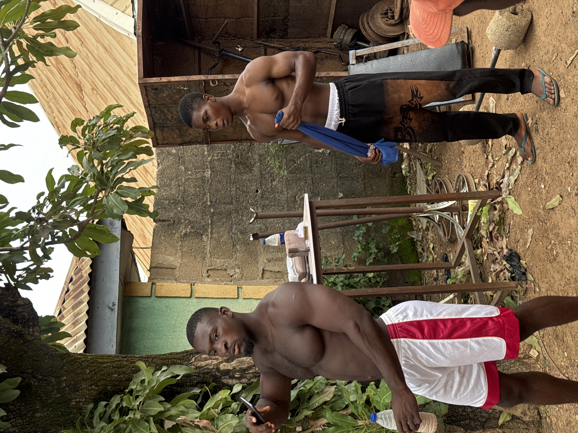 The image depicts an outdoor scene in Akwanga, Nigeria, possibly a gym or workout area. Two shirtless men are the central subjects. The man on the left, with dark skin and a beard, wears white shorts with red trim and holds a bottle of water in his left hand, and a smartphone in his right. The man on the right, also with dark skin, wears black athletic pants with brown markings and a white waistband, and holds a folded blue cloth in his right hand. They appear to be taking a break or preparing for a workout. The environment includes a large tree with green leaves on the left, a rustic building with a corrugated metal roof and a stone wall behind the men, and various workout-related objects scattered in the background, such as weights and metal equipment. The ground is covered with dirt and dry leaves, suggesting an outdoor, somewhat unrefined setting.
