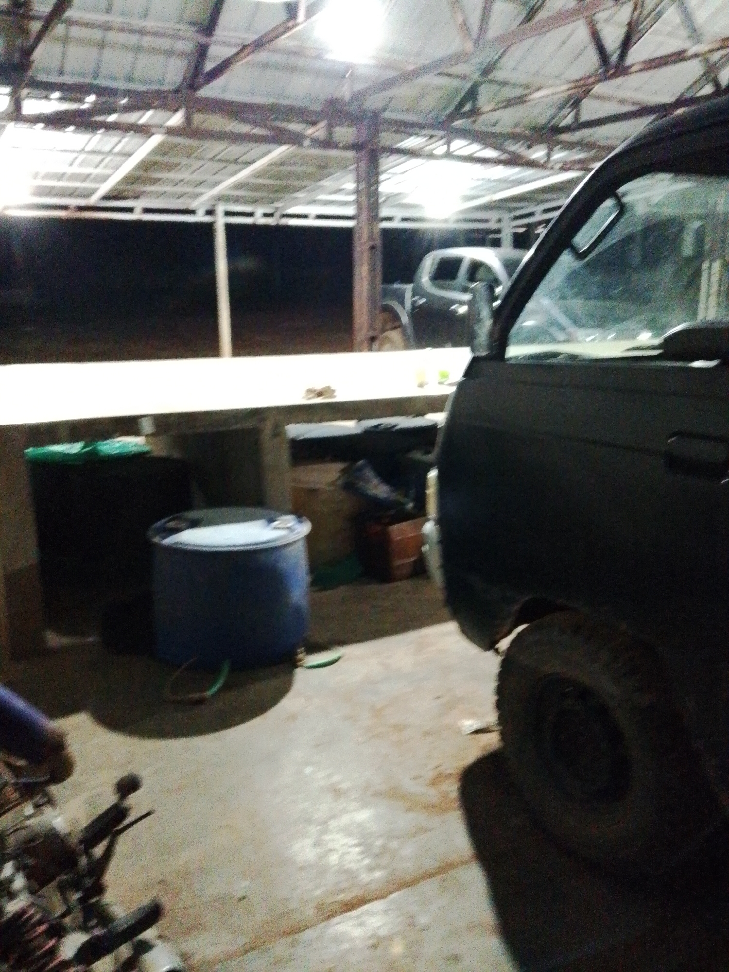 The photo appears to have been taken inside a metal-roofed structure, potentially a garage or shed, at night. Visible within are a dark-colored truck parked on the right side, two water barrels to the left, and what appears to be a white pickup truck parked further back in the shed. The location is likely Bukit Ketok, Indonesia. An engine of a motorcycle is in the foreground of the photograph.