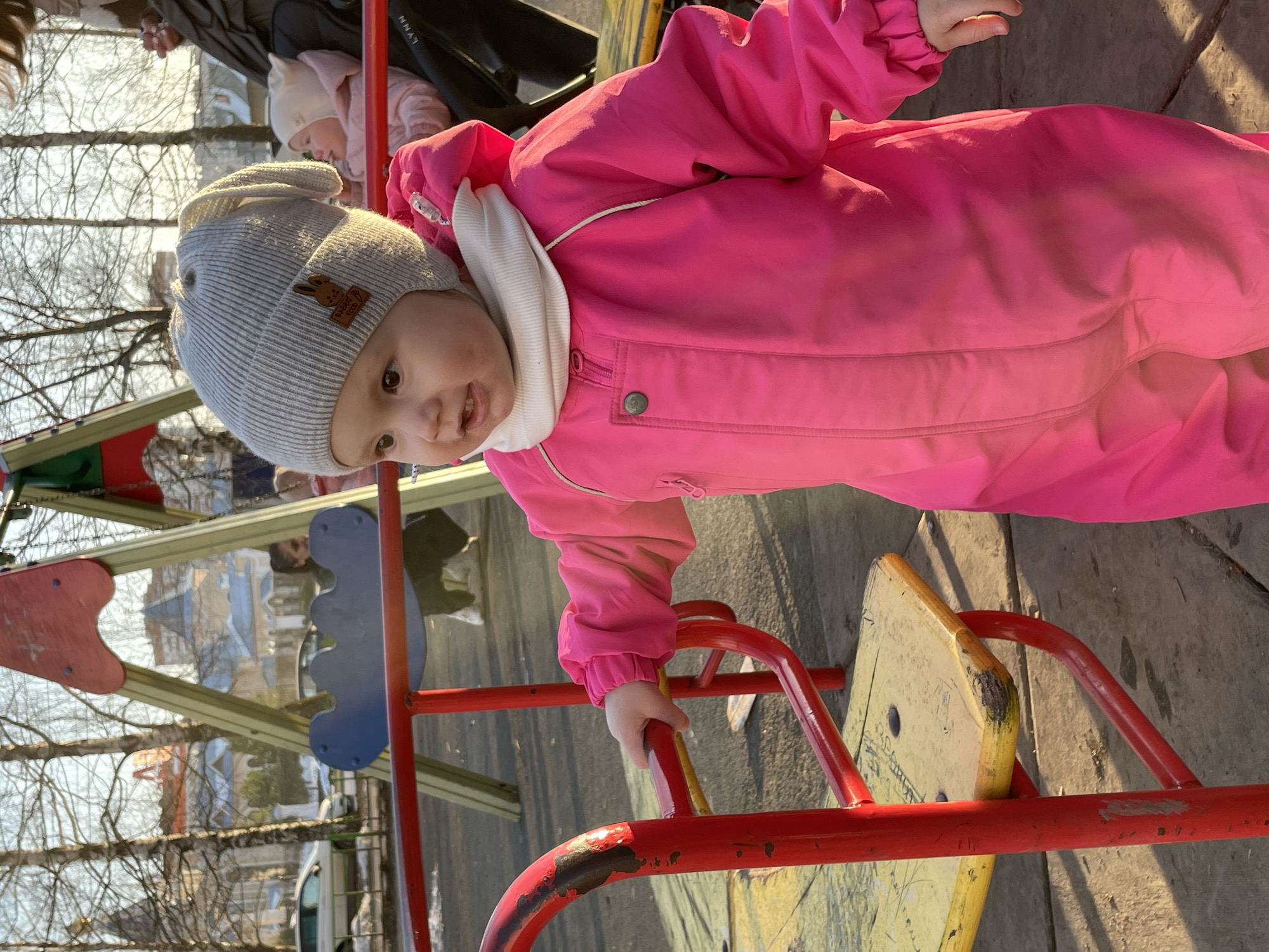 A toddler in a pink snowsuit and gray knit hat with a brown bunny patch is standing on playground equipment. The equipment has red metal bars and a yellow wooden seat. The toddler is holding onto the red bars with their left hand. Behind the toddler is a playground with blue and red elements, and further in the background are trees without leaves, reflecting sunlight. A stroller with another child is also visible in the background. The location is Izhevsk, Russia, as evidenced by the reflection in the playground elements and the overall architectural style of the surrounding buildings and landscape.