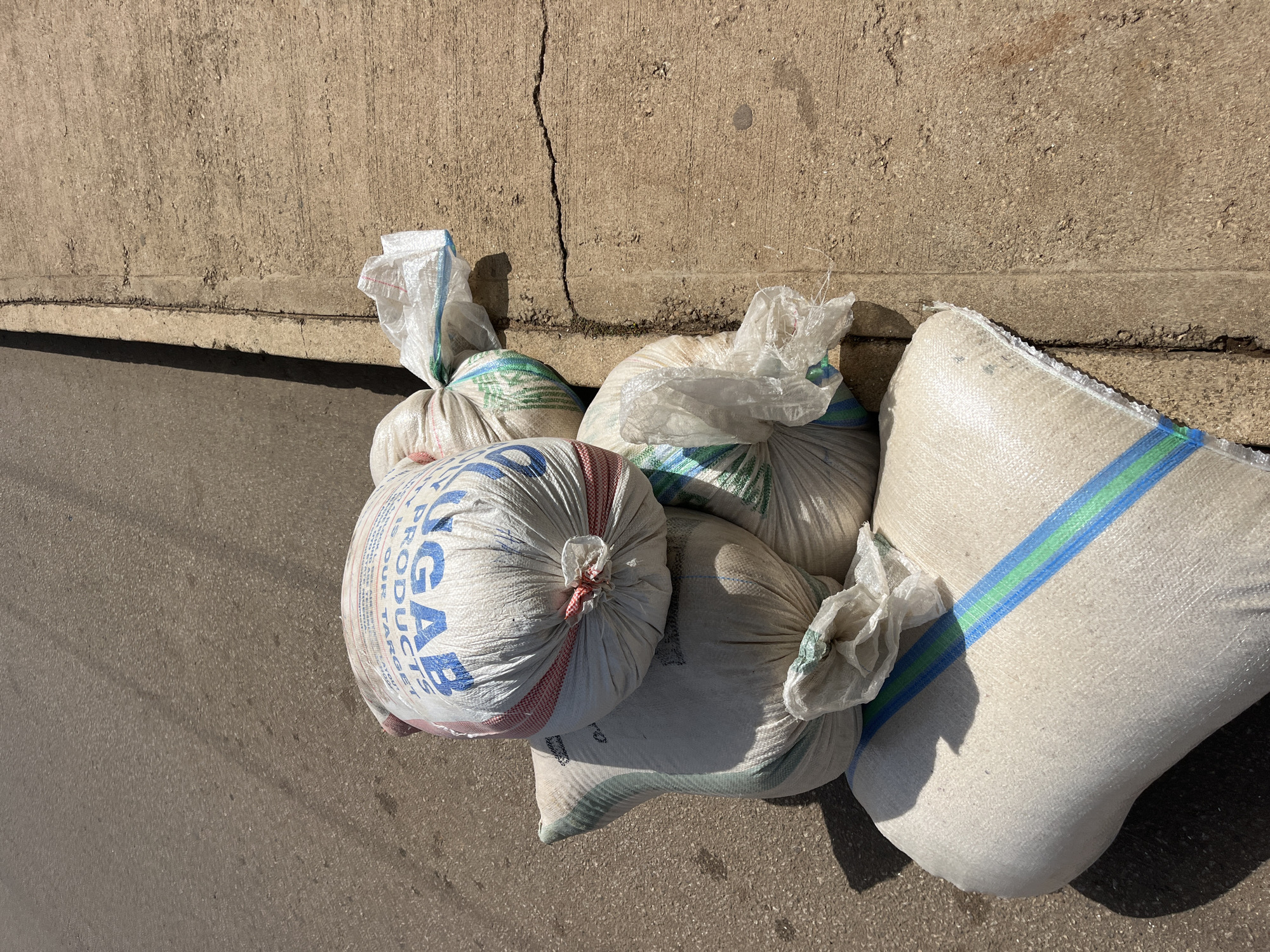 A pile of white woven sacks, some with blue and green stripes, are stacked on a cracked concrete sidewalk next to a paved road. One sack is partially visible with the word "SUGAR PRODUCTS" printed in blue on it. Sunlight casts shadows on the sacks and the road. There are no people visible in the image. The scene appears to be located in Bukuru, Nigeria.