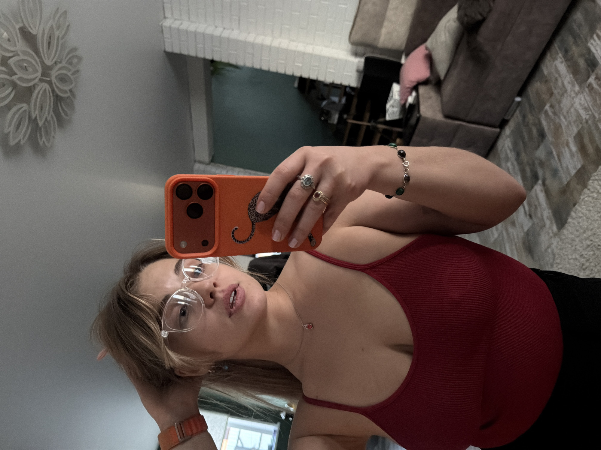 A woman is taking a selfie in an indoor setting. She is wearing a red tank top and clear-framed glasses. She has on a bracelet and rings. She's holding an orange phone with a case. The background includes a white brick structure, a glimpse of an exterior through an opening, a couch, and a wall decoration. The location is Большая Шильна, Russia.