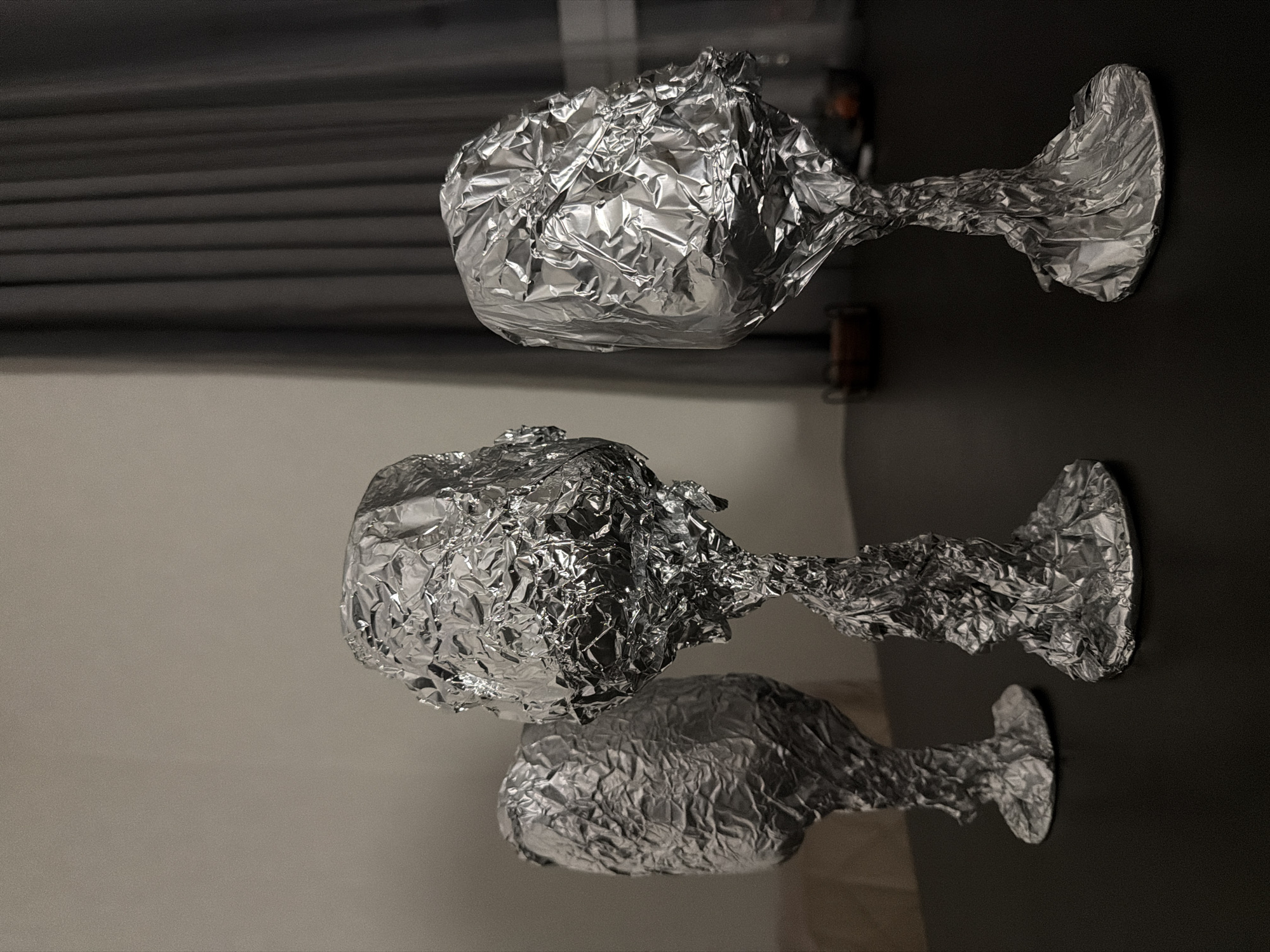 The image depicts three wine glasses completely wrapped in aluminum foil. The glasses are arranged on a surface, presumably a table. The background includes a gray curtain or wall and a white wall. The location is unknown.