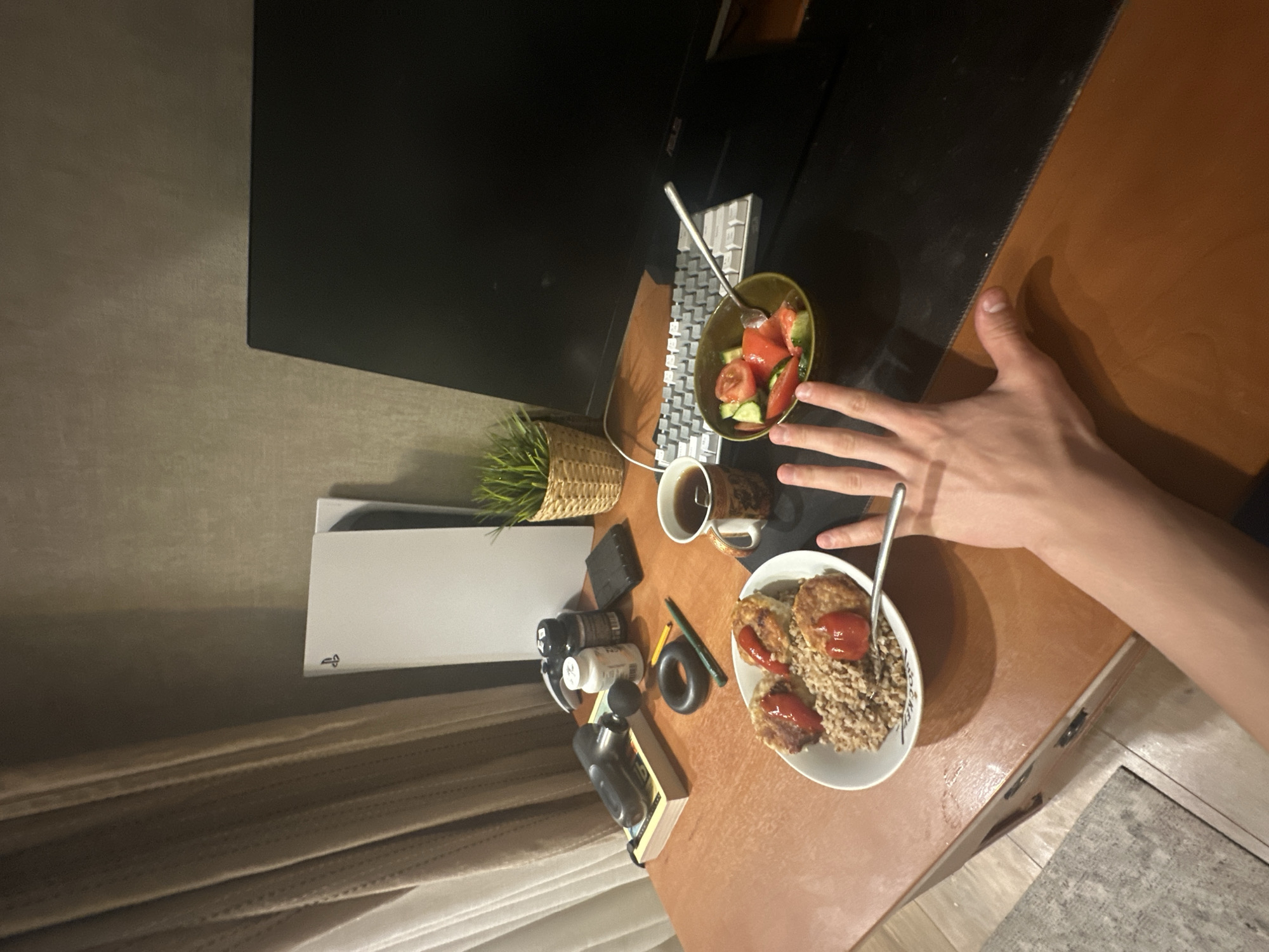 The image depicts a desk cluttered with various items. A computer monitor and keyboard are present, alongside a plant in a wicker basket. Food items are visible, including a salad in a bowl and a plate with what appears to be buckwheat and cutlets. A cup of coffee sits on the desk as well. Other objects include bottles, a book, a massage gun, and what seems to be a pen and ring. A hand is partially visible in the frame. A Playstation 5 console is placed next to the desk. The presence of a curtain suggests the desk is located indoors, possibly in a home office or study. No specific city or location within Unknown, Unknown can be determined from the image.