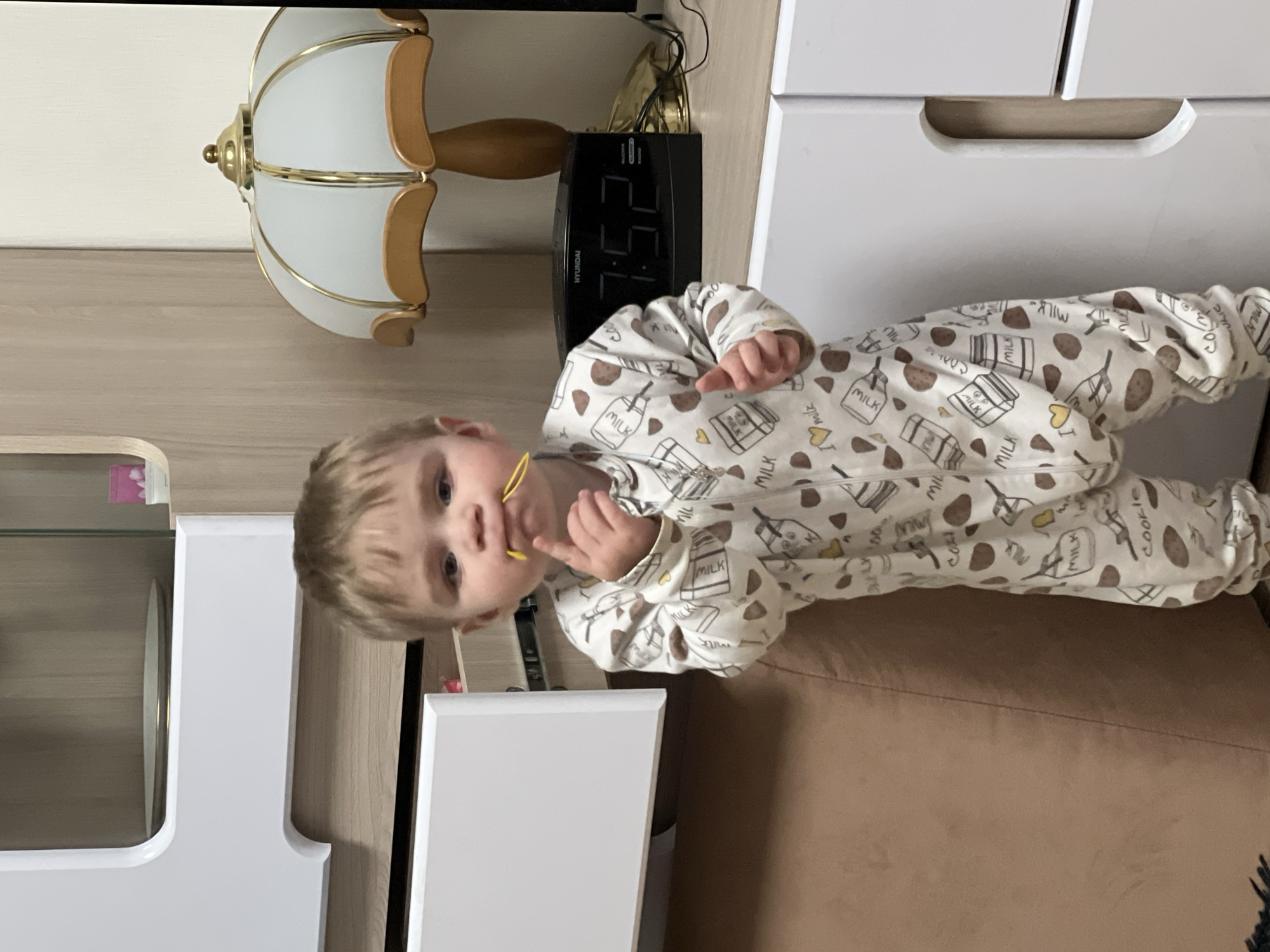 A young child wearing a white pajama with a milk and cookie pattern is looking at the camera. The child is standing next to a brown couch and a white shelf. A lamp and digital clock are visible in the background. The location is likely a home interior. There is no confirmation on the location being Perm, Russia.