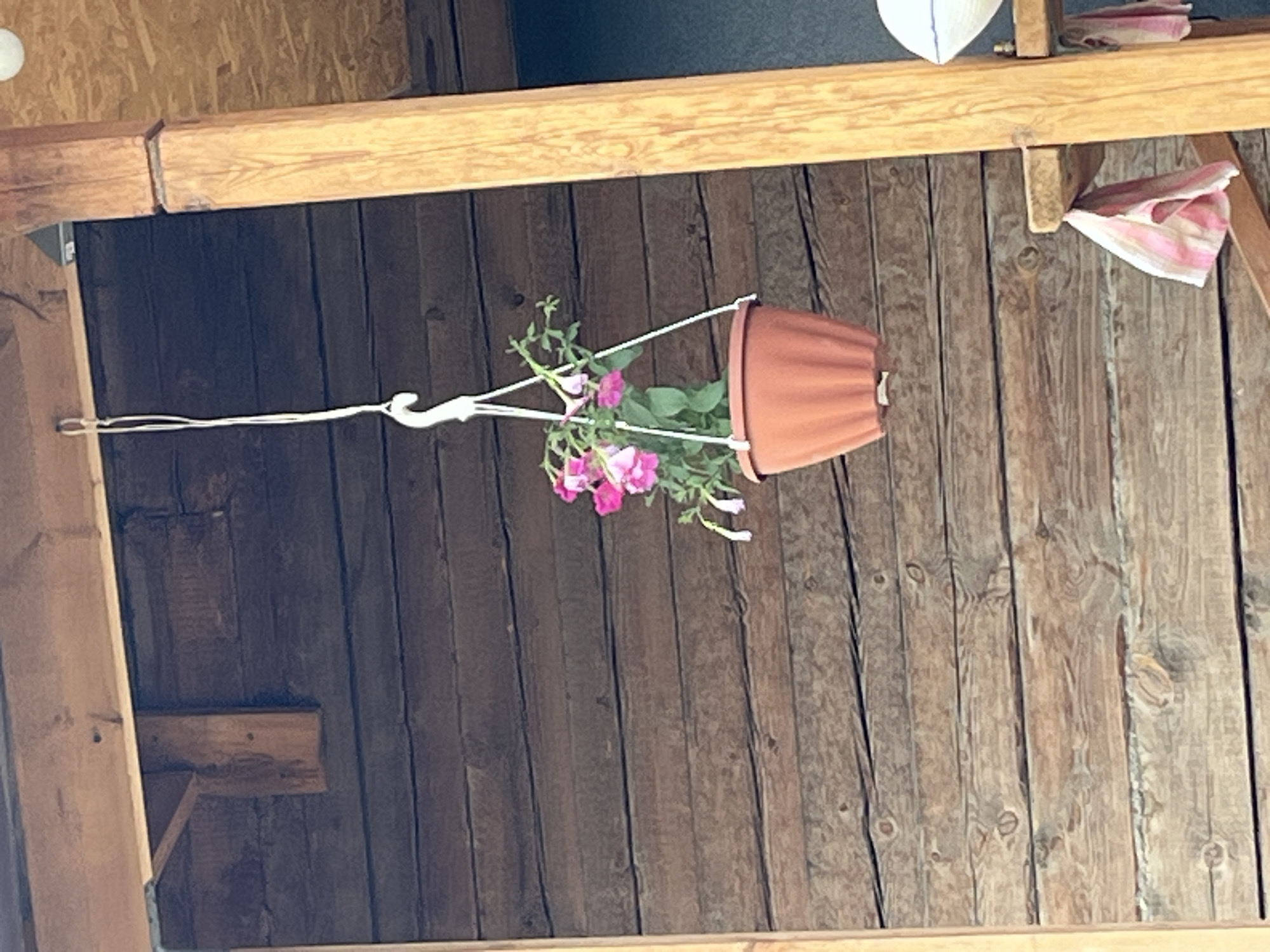 An overhead shot captures a hanging pot with pink flowers suspended from a white hook attached to a wooden beam. The pot, made of terracotta-colored plastic, is secured by a white cord. The background consists of horizontal wooden planks, creating a rustic texture. To the right, a light-colored wooden beam supports the structure. Partially visible at the bottom right is a shelf with two folded cloths, one pink and white striped, the other pink and white. The scene appears to be outdoors, possibly on a porch or under an overhang.