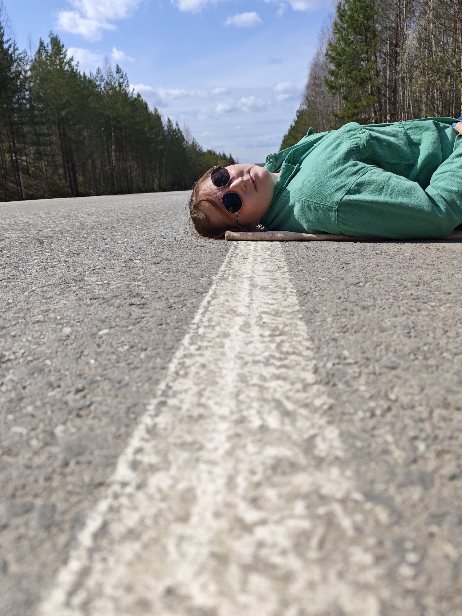 The image features a person lying on an asphalt road in Chaikovsky, Russia. The individual is dressed in a vibrant green jumpsuit or coverall and wears dark sunglasses, looking upwards towards the camera. They are positioned on their side, resting their head on what appears to be a light-colored fabric or mat.
The photograph is taken from a low-angle perspective, emphasizing the coarse texture of the asphalt road, which occupies the majority of the foreground and midground. A distinct white solid line, a road marking, runs longitudinally along the road surface, adding a linear element to the composition.
In the background, a dense forest of coniferous trees, likely pines, borders both sides of the road, suggesting a natural and possibly rural or remote setting. Above the treeline, a bright blue sky dotted with scattered white cumulus clouds indicates clear and sunny weather. The strong daylight illuminates the scene, highlighting the details of the person's attire and the surrounding environment.
There is no specific activity beyond the person lying on the road, possibly resting or posing for the photograph. No text is visible in the frame. The overall atmosphere appears tranquil and relaxed.