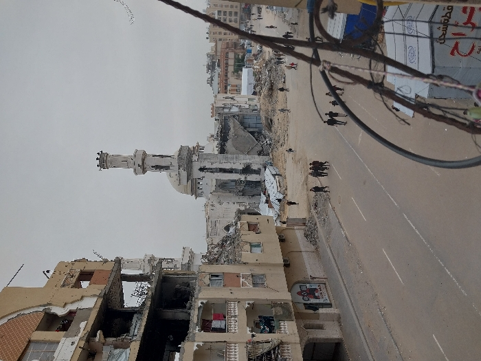 The image depicts a wide, elevated view of an urban landscape in Khan Yunis, Palestinian Territory, under an overcast sky. In the center midground, the Great Mosque of Khan Yunis stands heavily damaged, with large sections of its dome and main structure collapsed, and its minaret showing signs of compromise. The area immediately surrounding the mosque is covered in substantial debris and rubble. To the left, a multi-story residential or commercial building is extensively damaged, with several floors exposed, walls missing, and structural wreckage visible. Clothing is hanging from balconies on the lower floors of this building. A mural is present on its ground floor facade.
A paved street runs horizontally across the midground, separating the foreground from the mosque and other structures. The street features double white lines in its center and a single white line along the curb. Debris and smaller rubble piles line the edges of the street. Numerous individuals and small groups, totaling approximately 10-15 people, are observed walking on the street and traversing the rubble-covered ground between the road and the mosque. Some people are also visible further in the background amidst the destruction.
The immediate foreground on the right is partially obscured by a tangle of dark electrical wires and cables. Below these wires, a white tent-like structure with red and black Arabic script is partially visible. In the background, beyond the mosque, more damaged buildings and rubble extend into the distance, with some less damaged structures discernible further back. A flock of birds is visible flying in the upper right quadrant of the gray, featureless sky.
