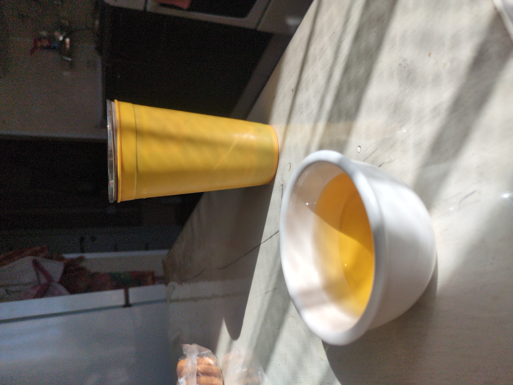 The image shows a table with a white bowl containing a yellow liquid, a yellow cup, and a partially visible plastic bag containing some round objects, possibly cookies. The table appears to be made of a light-colored material with a subtle pattern. Sunlight casts strong shadows across the table, indicating a bright, sunny environment. In the background, a dark countertop and a portion of a kitchen area are visible. To the left, a door is slightly ajar, revealing a glimpse of what appears to be a person in patterned clothing standing in a doorway. The overall setting suggests an indoor scene, likely a kitchen or dining area. The image was taken in the city of Kulebaki, Russia.