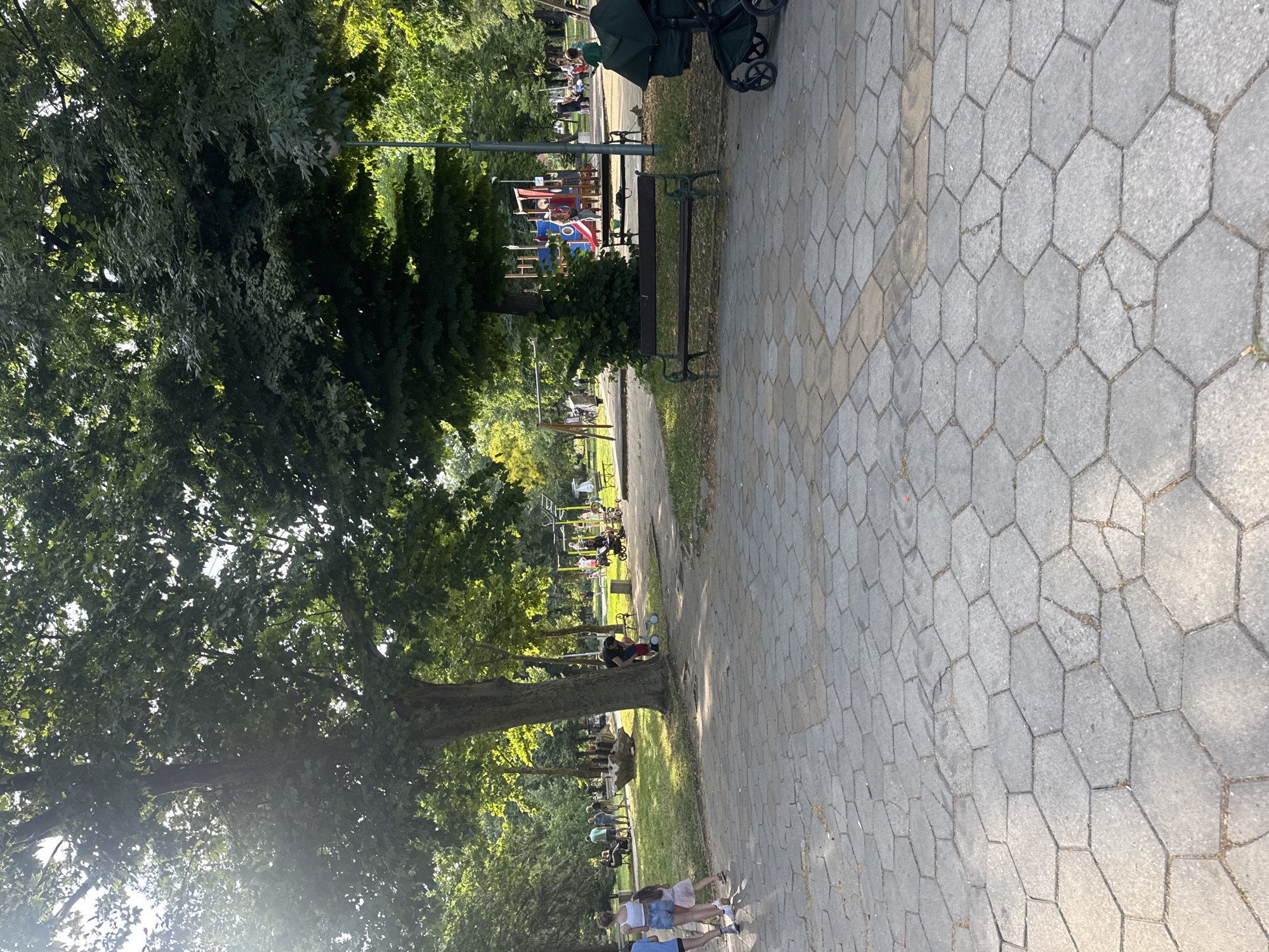 The image depicts a park scene in Plovdiv, Bulgaria. In the foreground, a paved area with hexagonal stones leads into a grassy expanse dotted with trees. Several people are scattered throughout the park. On the left, three young girls are walking on the paved area. Further back, a group of adults are gathered, some sitting on benches. In the middle ground, a playground area with swings and climbing structures is visible. A man is seen bending down near a small child on a tricycle. To the right, a dark green stroller is parked next to a wooden bench. The environment is lush and green, suggesting a warm, sunny day, with the sun's rays filtering through the tree canopy.