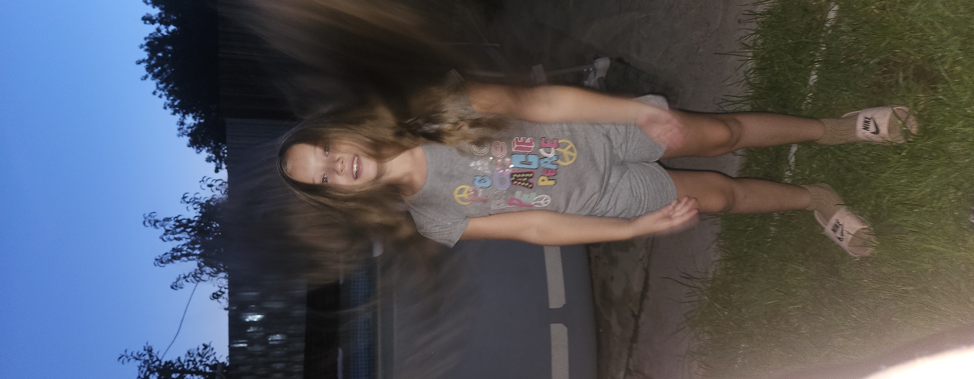 A young female, with long blonde hair appearing blurred due to motion, stands in a grassy area at dusk. She is wearing a grey t-shirt with colorful lettering and peace symbols, and grey shorts. Her feet are clad in light-colored slides with the Nike logo. The background shows a blurred swimming pool and a fence. The sky is a deep blue, indicative of twilight. The location is identified as Semey, Kazakhstan.