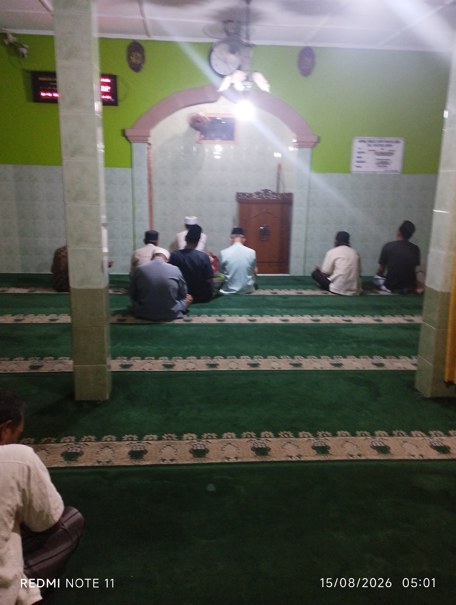 This image depicts the interior of a mosque in Karawang, Indonesia. The scene is captured from a low angle, with the foreground dominated by a person kneeling on a green carpet adorned with patterned borders. In the midground, several men are seen sitting on the carpet, facing away from the camera, some wearing prayer caps. The background reveals a mosque interior with light green walls, an arched doorway, and a wooden pulpit. Two digital clocks are visible on the wall. The overall lighting suggests an indoor setting, likely during a prayer time or religious gathering. The timestamp at the bottom indicates the image was taken on August 15, 2026, at 05:01.