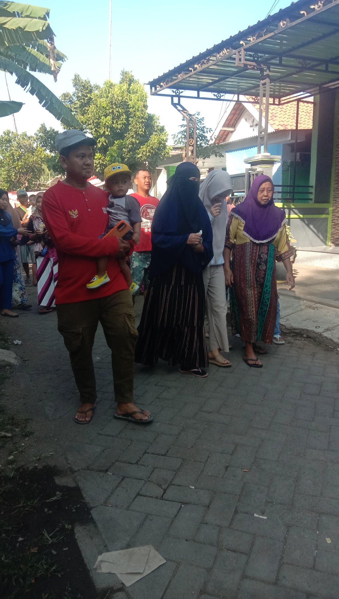 In Ngempit, Indonesia, a group of people are gathered outdoors on a paved pathway. The focus of the image is a man in a red long-sleeved shirt and olive-green cargo pants, holding a young child wearing a yellow cap. The man is wearing a grey baseball cap and dark flip-flops. To his right, a child in a grey shirt with a yellow cap is being held. Behind them, several other individuals are visible. A woman in a dark hijab and a striped skirt walks with a younger woman in a light grey hijab and light-colored pants. Further to the right, an older woman in a colorful dress and purple hijab is also present. The environment includes trees, a building with a covered awning, and a paved ground with some scattered debris. The sun is shining, casting shadows.