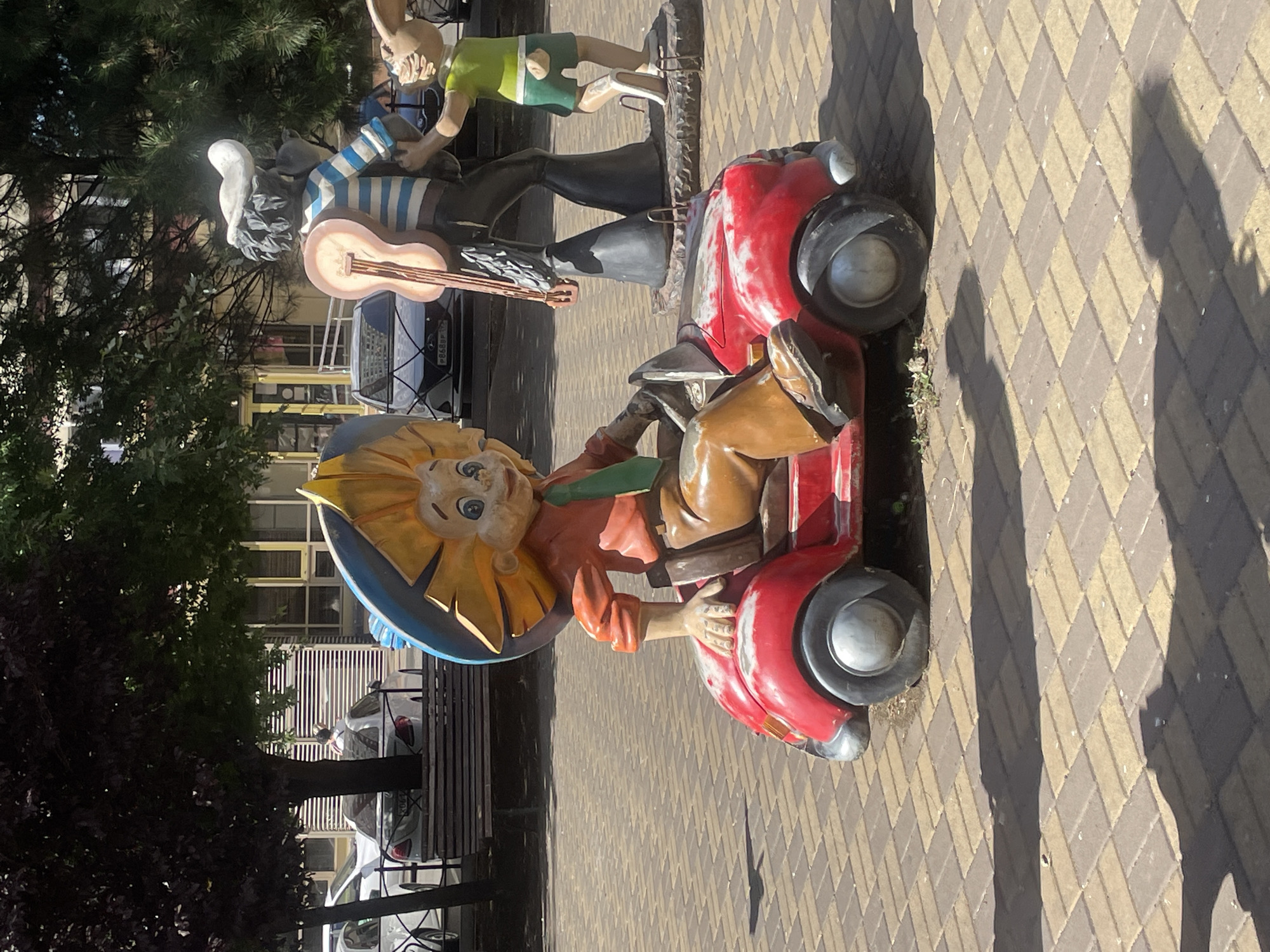 This image depicts a whimsical outdoor sculpture scene. In the foreground, a cartoonish character with a large, yellow sun-like hat, orange shirt, green tie, and brown pants is seated in a small, red toy car. The sculpture has a playful expression. Behind this, another sculpture features a character in a blue and white striped shirt, dark pants, and a white cap, holding a guitar. To the right, a sculpture of a rabbit wearing a green shirt and shorts stands. The sculptures are situated on a paved area with a repeating pattern of brown and beige rectangular tiles. In the background, trees, buildings with windows, and parked cars are visible. The scene appears to be located in a public outdoor space, possibly a park or a city square. The location is identified as Krasnodar, Russia.