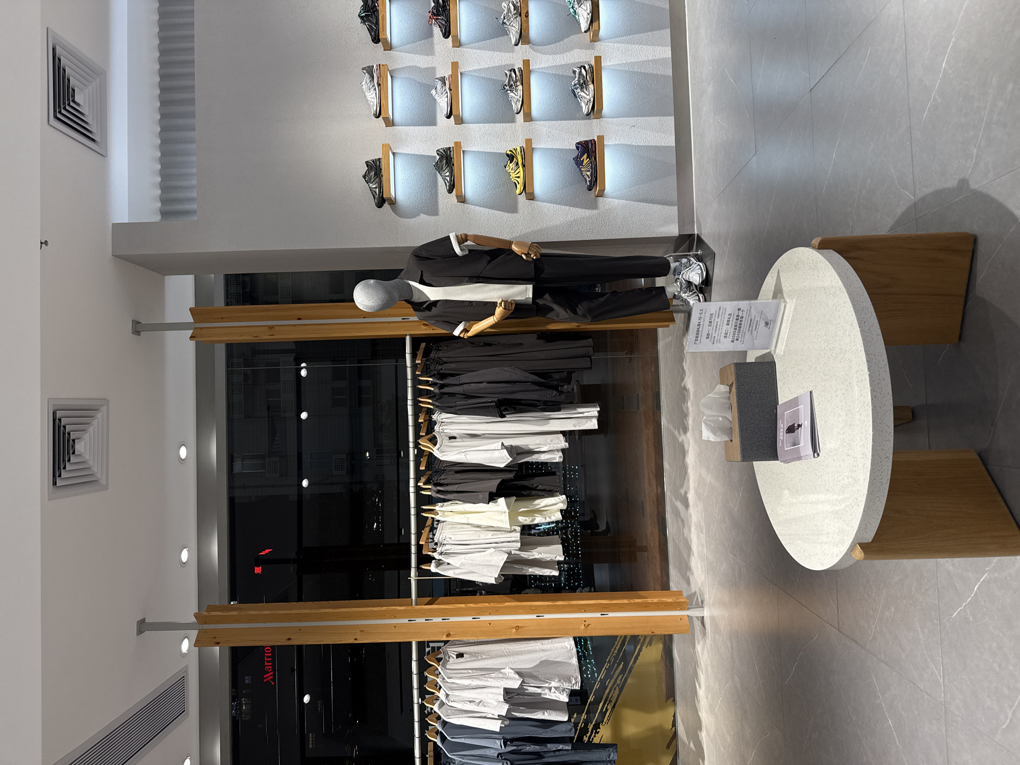 The image shows the interior of a retail clothing store, possibly a branch of a chain like Marriott, as indicated by a sign visible through the window behind the clothing racks. The location appears to be in the Tianhe District of China, based on the presence of Chinese characters and the sign promoting the China Import and Export Fair (广交会).
A mannequin is positioned against the back wall, dressed in a black suit with a white undershirt. Above the mannequin, numerous pairs of sneakers are displayed on individual shelves.
Racks of clothing, mostly white and grey garments, are arranged behind a glass partition with wooden framing. A round table with two stools sits in the foreground. The table holds a tissue box and informational pamphlets in front of it. The floor is tiled in grey. The light fixtures include square air vents and recessed lighting on the wall and ceiling.