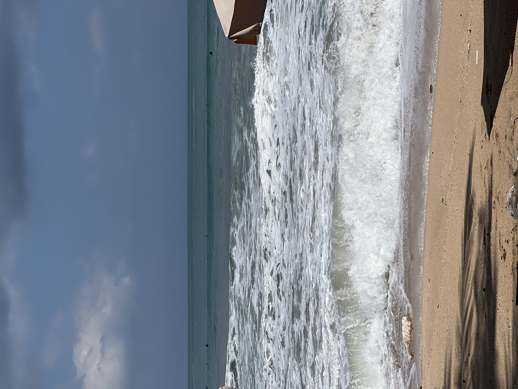 The image captures a beach scene in Kuta, Indonesia, featuring a sandy shore in the foreground with scattered debris and the shadow of palm leaves. The ocean is active with white-capped waves rolling towards the beach. Several surfers can be seen in the middle distance, appearing as small dark figures in the water, some with surfboards. The water transitions from turquoise to a deeper blue towards the horizon, where it meets a grey, overcast sky. On the right side of the frame, a portion of a light-colored beach umbrella or canopy is visible.