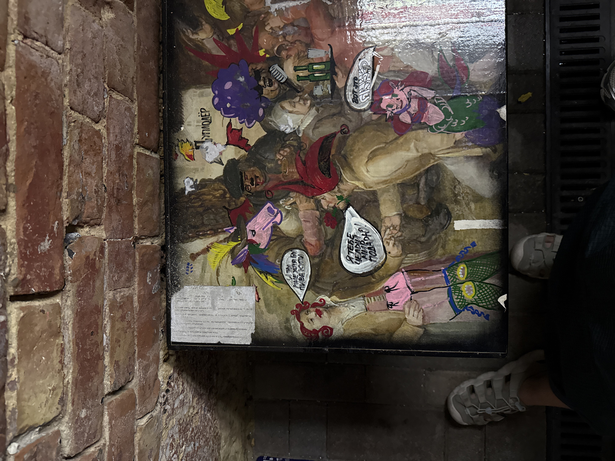 This image shows a section of a brick wall and a table with a painting on it. The painting depicts a lively scene with multiple figures in what appears to be a tavern or gathering. The figures are rendered in a somewhat cartoonish style, with exaggerated features and bright colors. Some figures have speech bubbles containing text in Cyrillic script. There are also cutouts of figures applied to the painting. The table is positioned against the brick wall. The lower part of the image shows a person's feet wearing sneakers, standing near the table. The environment appears to be indoors, possibly in a historical or rustic setting, given the brickwork. The painting likely contains elements of Russian culture or humor. There is no explicit information in the image to pinpoint the exact location in Rostov-on-Don, Russia.