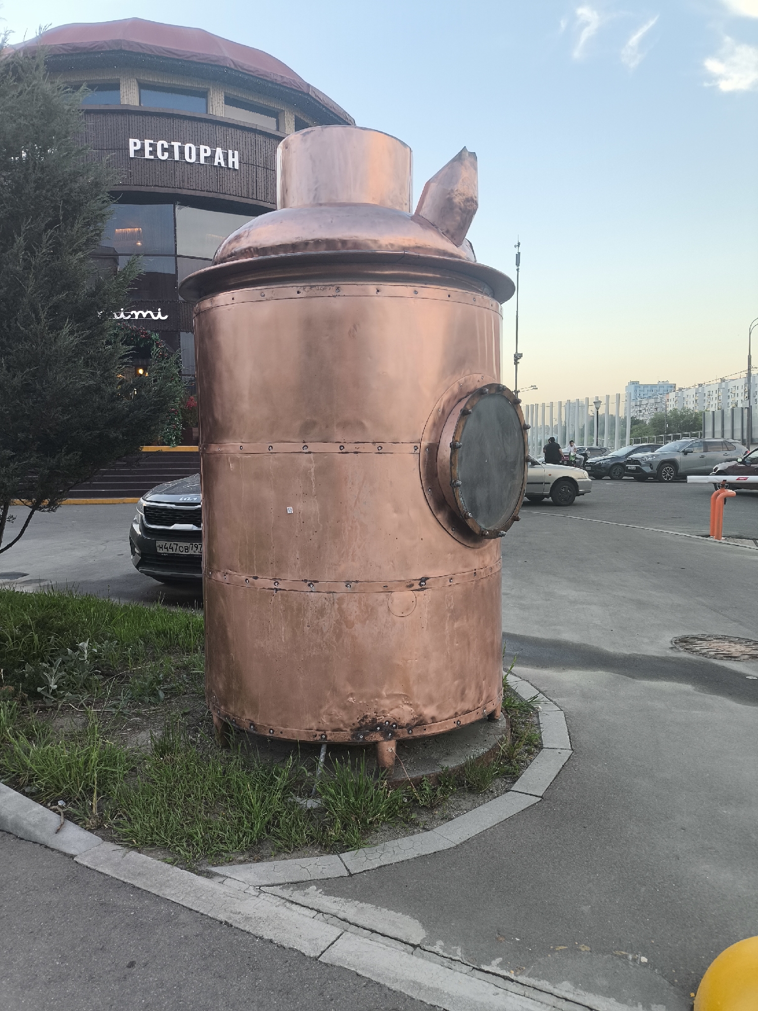 The image depicts a large, copper-colored metal structure resembling a stylized distillation apparatus, situated on a grassy patch adjacent to a paved area. It has a cylindrical body with bolted seams, a circular viewing port, and a bulbous top with angled attachments. Behind the structure, a modern building with the word "РЕСТОРАН" (restaurant) in Cyrillic script is visible. A gray Kia Seltos SUV is partially obscured by the copper structure, while other cars are parked in the background. Several people are visible near the cars. The scene is set in daylight under a clear sky, suggesting an urban location.