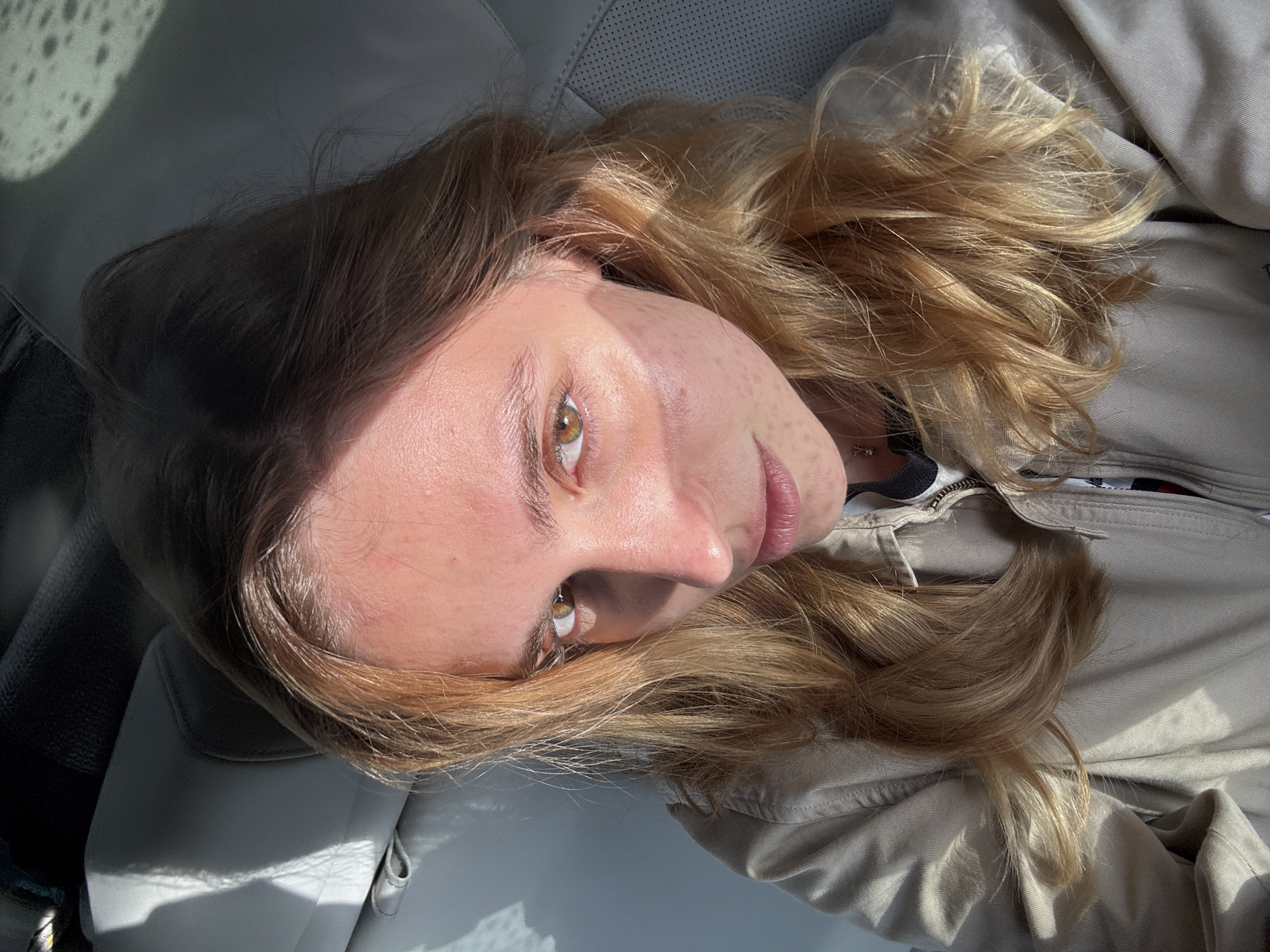 A young woman with fair skin and light brown hair is lying down, possibly in a car, as indicated by the white leather-like surface beneath her. She has hazel eyes and freckles on her forehead. She wears a light-colored jacket with a visible zipper. Sunlight is streaming in, creating shadows and highlights across her face and the surrounding surfaces. The location is not discernible from the image.