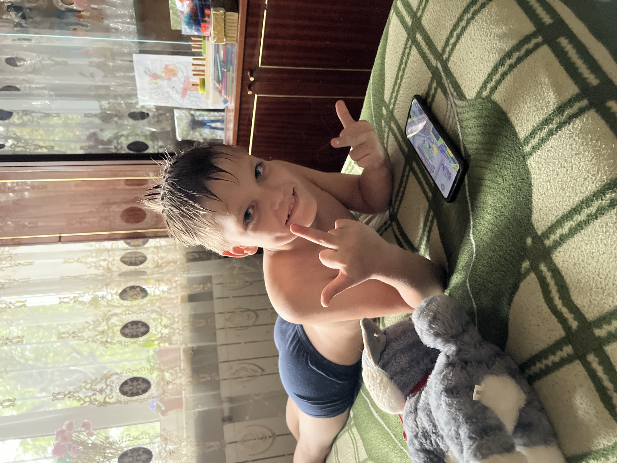 A young boy, likely around 5-7 years old, lies on a bed covered with a green and white checkered blanket. He is looking up at the camera and smiling, with his right hand raised and making a peace sign, while his left hand is also raised with fingers pointing up. He is wearing dark blue boxer briefs. To his right, a smartphone displays a game or application with colorful graphics. A grey stuffed animal with white accents is nestled next to him on the bed.
In the background, a wooden cabinet with dark brown doors is visible. On top of the cabinet, there are various items including what appear to be art supplies like colored pencils and a coloring book page depicting a cartoon character. Lace curtains hang in front of a window, allowing natural light to illuminate the scene. Through the curtains, greenery from trees is visible, suggesting it is daytime and potentially a warm season. The overall mood appears relaxed and playful. No visible text is present in the image. The setting is likely a bedroom within a residential space in Qarağandı city administration, Kazakhstan.