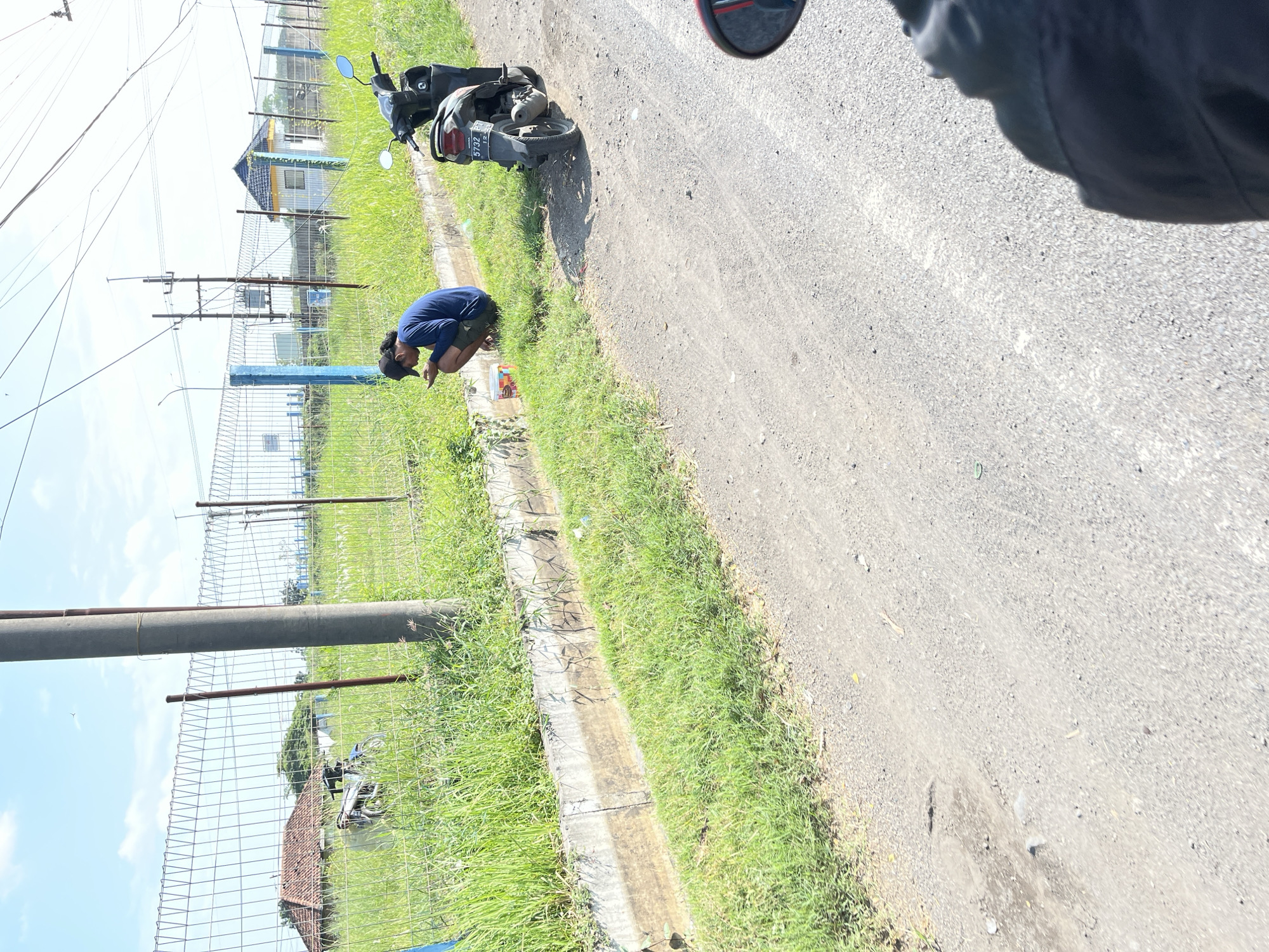 A young man with dark hair and a black cap, wearing a blue long-sleeved shirt and dark shorts, is crouching on the grassy shoulder of a dirt road. He is looking at his phone, which he holds in his hands. Next to him, a red and white bucket is on the ground. A dark-colored motorcycle is parked on the road to his right. In the background, a chain-link fence runs along the edge of a field of tall green grass. Beyond the fence, there is a building with a yellow roof and a smaller white structure, as well as a house with a red tiled roof. A large concrete utility pole stands on the left side of the frame. The sky is clear and blue with some scattered clouds. The scene appears to be located in a rural or semi-rural area in Pamotan, Indonesia, given the context. The lighting suggests it is daytime, likely late morning or afternoon, due to the strong shadows.