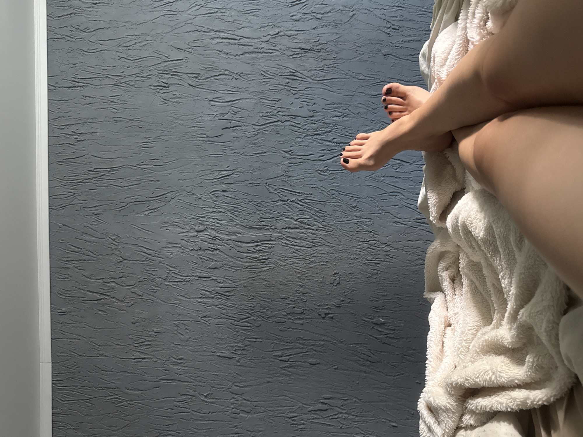 The image depicts a close-up view of a person's legs and feet resting on a plush, cream-colored blanket. The background is a textured gray wall with vertical striations, and a white crown molding runs along the top edge. The lighting suggests a natural light source from the side, casting shadows on the wall and highlighting the texture of the blanket and the skin of the legs. The toenails are painted black. The scene appears to be indoors, likely a bedroom, and does not contain any specific elements that would indicate its location within the city of Aznakayevo, Russia.