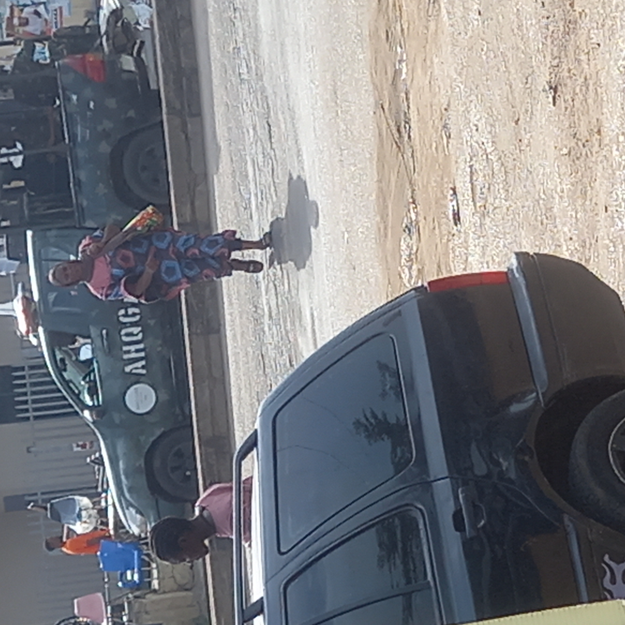 An eye-level shot presents a scene on what appears to be a paved street in Dadi Riba, Nigeria. A Black SUV takes up a significant portion of the foreground, its rear and side visible to the right. To the left of the SUV, an individual with a pink shirt stands facing forward.
Further into the shot, an older woman wearing a patterned pink and blue dress and holding an unidentified colorful item, stands in the street. Adjacent to her is a green vehicle with "AHOG" printed on the side in white capital letters, in vertical format.
In the background, other individuals and vehicles are present, creating a sense of an urban environment. The ground exhibits a light tan, textured surface and an elevated wall can be seen on the left edge of the image.