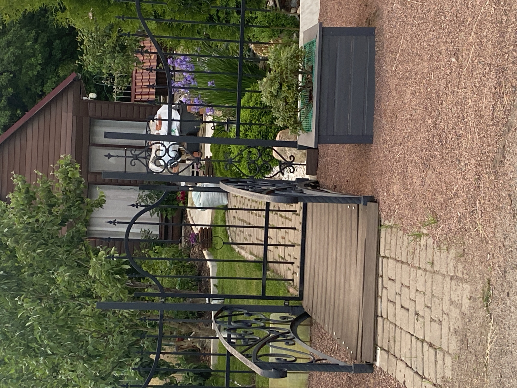 The image shows a garden scene in Bol'shoe Zaborod'e, Russia, featuring a wooden footbridge with ornate metal railings leading over a paved pathway. The pathway is made of brick pavers and is surrounded by reddish-brown gravel. To the right of the bridge, a dark gray planter box contains a small, bushy green plant. In the background, behind a black metal fence, a brown wooden structure, possibly a shed or small house, is visible with windows and a red-tiled roof. Several green trees and bushes are present, along with some purple flowers. The overall impression is of a well-maintained, private garden.