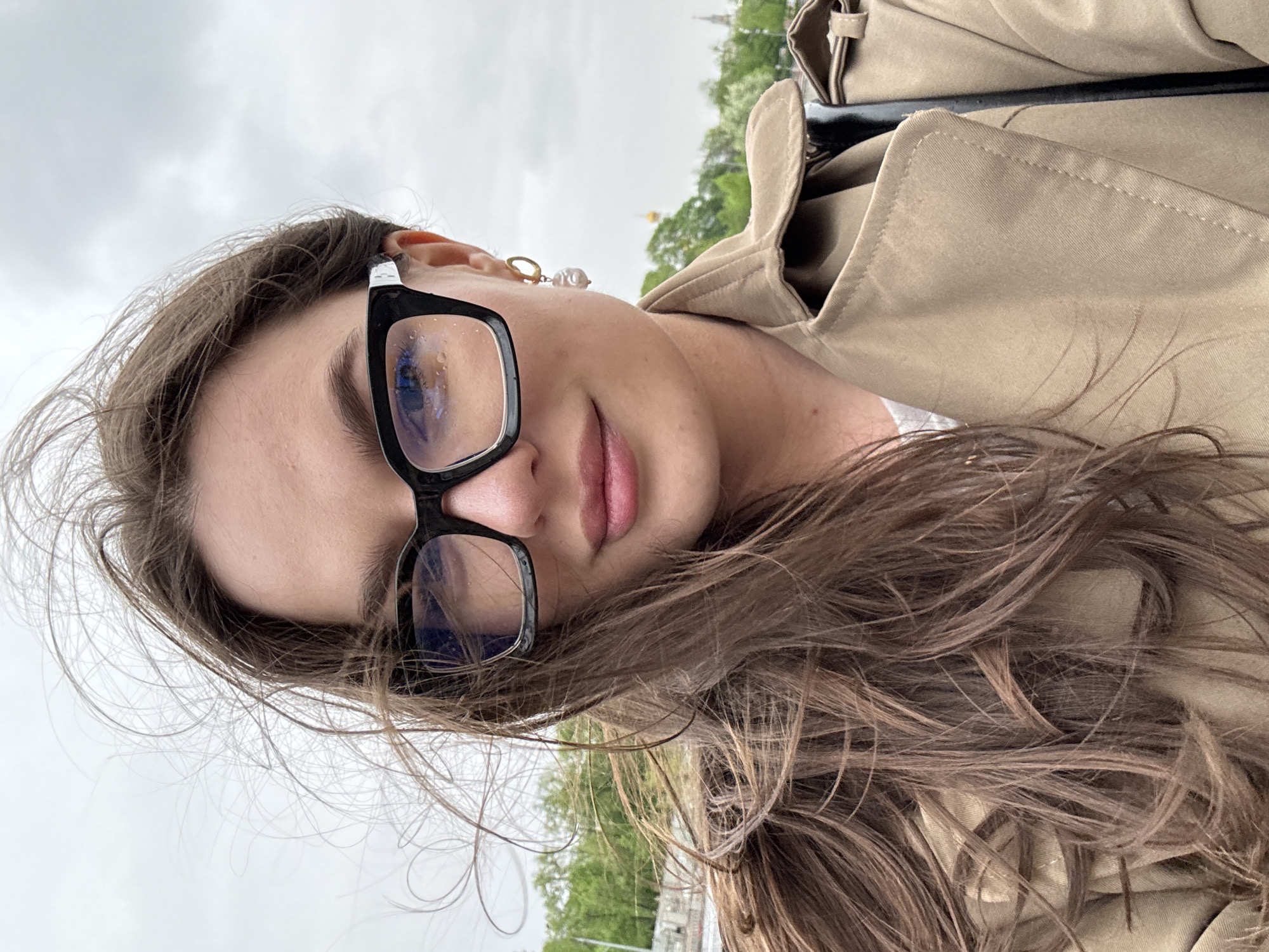 A woman is pictured in a close-up selfie, likely in an outdoor urban setting. She has light skin, brown hair, and wears black-framed glasses. She also has a gold hoop earring with a pearl attached. She is wearing a tan coat or jacket. The background appears to be outdoors with trees and a pale, overcast sky. A distinct building with a golden dome is visible in the background, suggesting the location is Moscow, Russia.