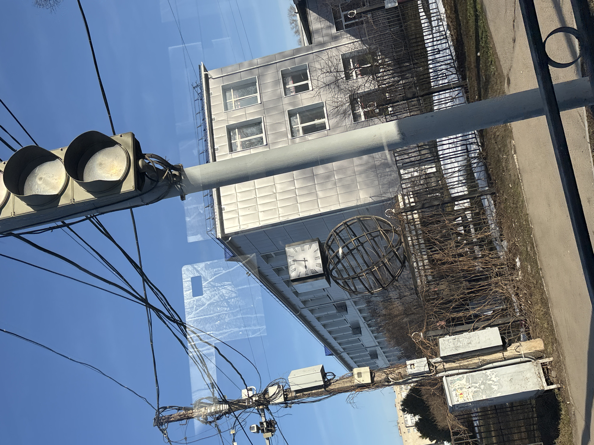 The image shows a view through a window in Chaikovsky, Russia. Reflections in the window overlay the scene outside. A gray building with square windows is visible. A clock is mounted on the building's facade next to a metal spherical structure. Below the building, a utility pole laden with wires and junction boxes is present. To the left, a traffic light fixture with two lights is affixed to a pole. The sky is blue. A fence and patches of snow are seen along the side of the road.