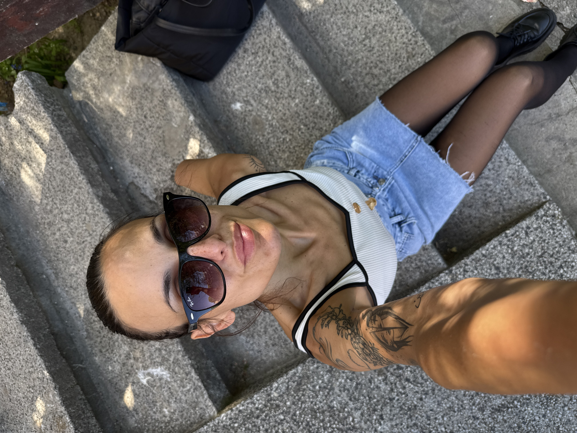 A young woman is the central subject of this high-angle photograph, taken from directly above as she lies on her back on a set of outdoor concrete steps. She is wearing large dark sunglasses, a white and black ribbed tank top, distressed blue denim shorts, sheer black tights, and black lace-up shoes. Her dark hair is pulled back, and she has several tattoos, prominently featuring what appears to be a nautical or ship design on her left forearm, as well as a smaller symbol on her right arm/shoulder. A black backpack or bag is visible on the steps above her head.
The activity taking place involves the woman posing for the camera, looking upwards. Her left arm is extended towards the lens, suggesting she is taking a selfie or being photographed by someone standing directly over her. She appears relaxed in her pose.
The scene is brightly illuminated by natural daylight, indicated by strong shadows cast across the rough, grey concrete steps. Some green foliage is visible along the left edge of the frame, suggesting an outdoor setting. The word "Ray-Ban" is visible on the right lens of her sunglasses. The image was taken in Pleven, Bulgaria.