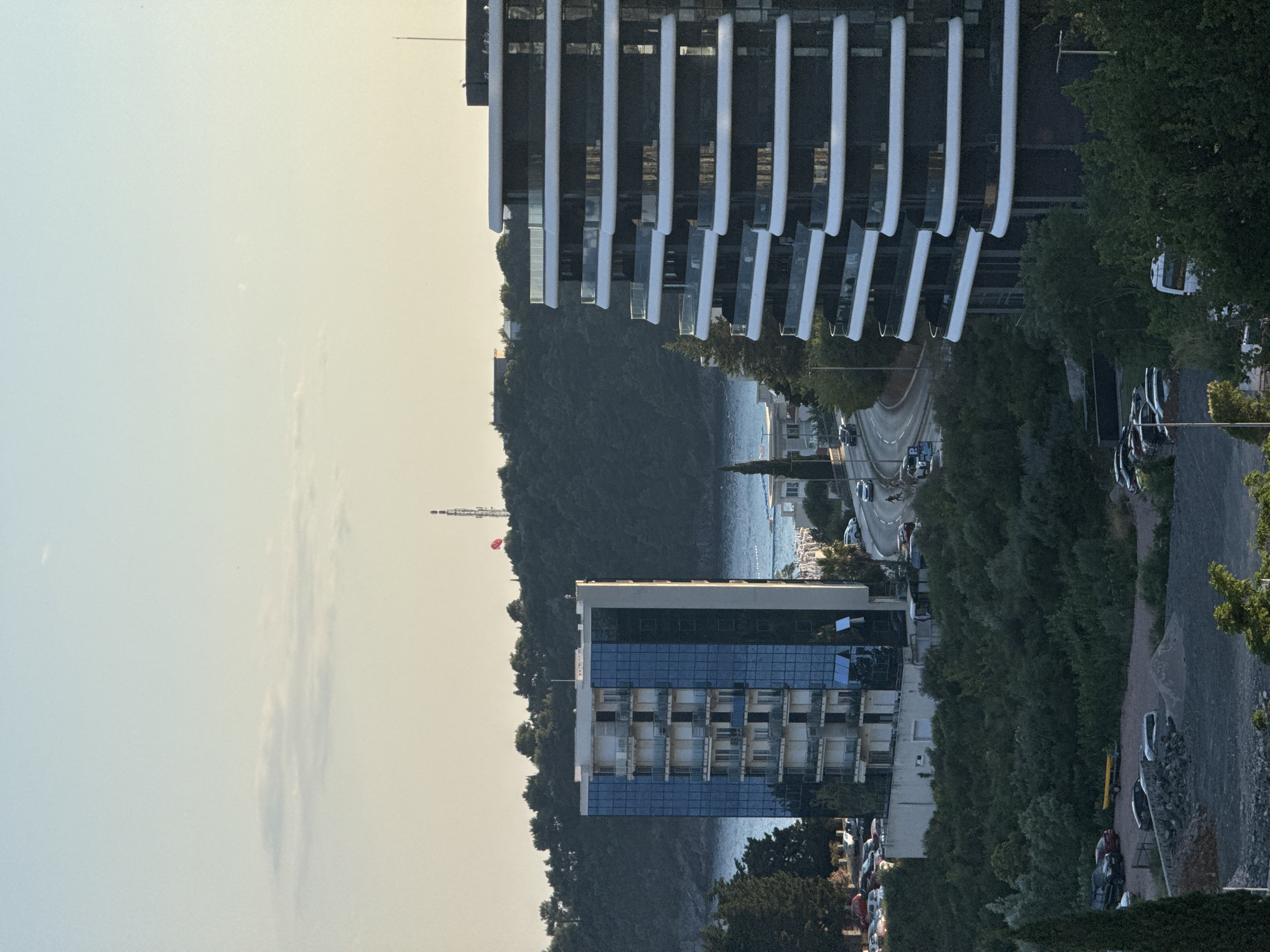 The image captures a scenic view of the city of Boreti, Montenegro, at the golden hour. In the foreground, a modern, multi-story building with a dark facade and white, curved balconies stands prominently. To its left, another building with a glass facade and white balconies is visible. A road winds its way through the scene, with a few cars and pedestrians. Lush green trees and vegetation cover the landscape, leading down to the sparkling blue sea. In the distance, a red parasail is suspended in the air, adding a touch of dynamism to the otherwise serene environment. The sky is a gradient of soft blues and oranges, with a few wispy clouds. The overall impression is one of a tranquil coastal city bathed in the warm glow of the setting sun.