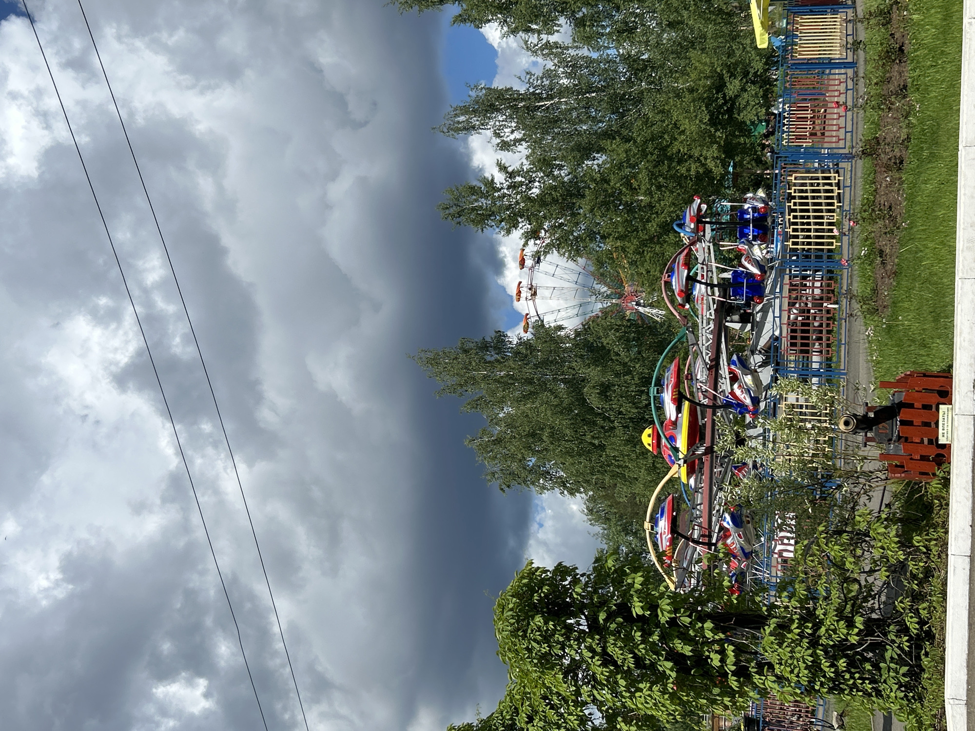 A Ferris wheel and spinning amusement ride are situated among trees in what appears to be an amusement park. Overcast skies indicate possible inclement weather. Electrical wires are seen in the upper-left of the frame. The location is most likely in the city of Chaikovsky, Russia. No people are visible.