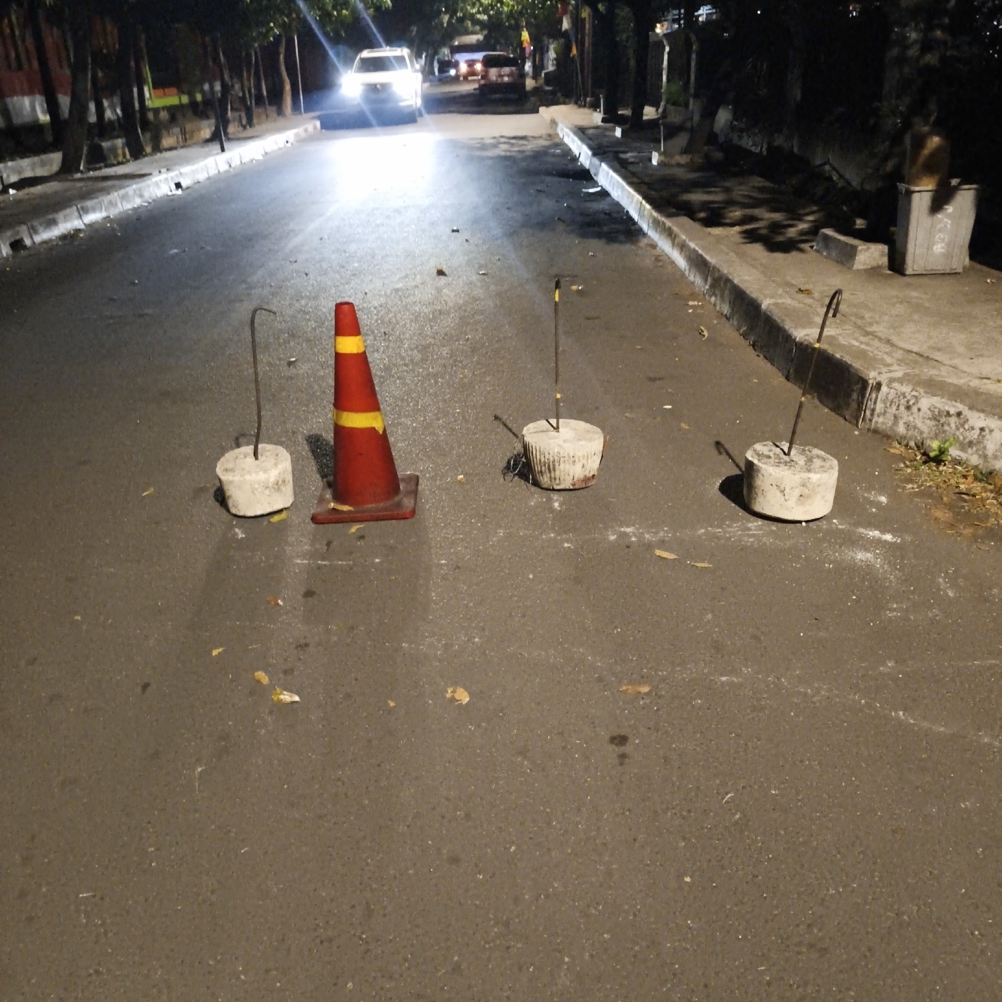 The image depicts a street scene at night in South Jakarta, Indonesia, likely on Jalan Ampera Raya. The asphalt road shows signs of wear and tear, with scattered debris including leaves and what appears to be white powder. On the right side of the road, a concrete curb separates the road from a sidewalk. A gray trash bin with some writing on it is visible on the sidewalk.
In the foreground, a red and yellow traffic cone stands prominently. To its left and right are three cylindrical concrete blocks, each with a dark metal rod protruding vertically from its center. These concrete blocks appear to be anchors or weights, possibly for temporary barriers or marking.
Further down the road, several vehicles are illuminated by their headlights, indicating traffic. A white car is the most visible, with its headlights casting a strong glow onto the road. Other vehicles, including what appears to be a red car, are also present. Trees line the left side of the road, with their leaves forming a dark canopy. The overall atmosphere suggests a typical urban street at night, potentially with some ongoing roadwork or maintenance indicated by the traffic cone and concrete anchors.