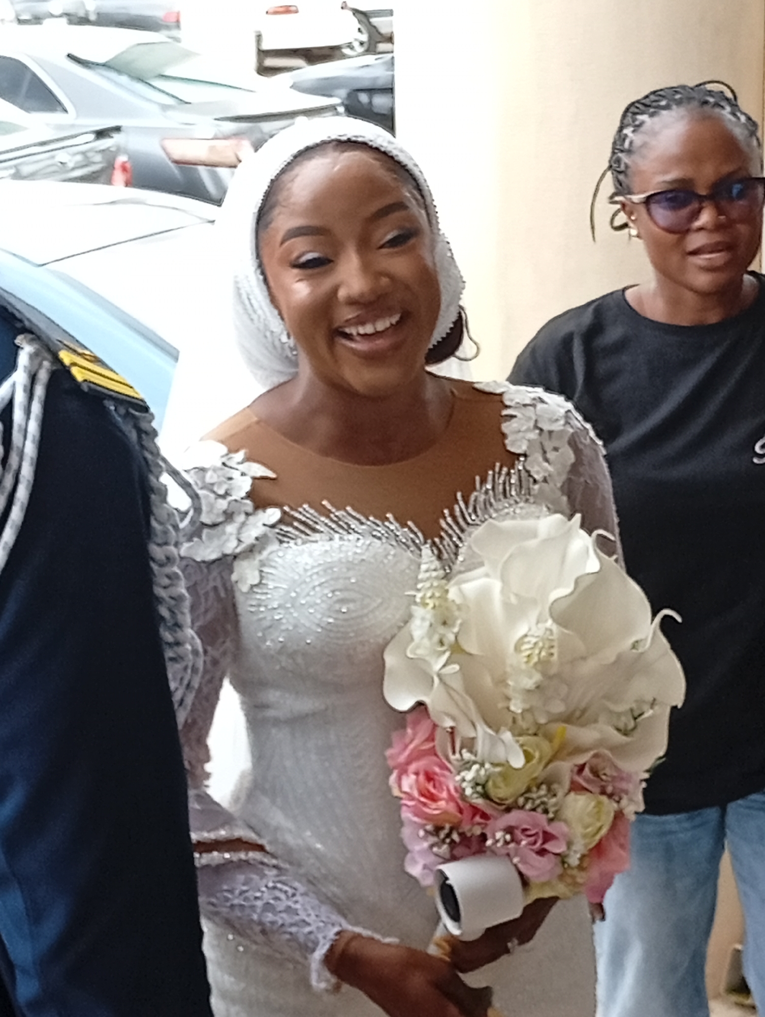 In Barden Yana, Nigeria, a newlywed woman in a white wedding dress and headscarf smiles, holding a bouquet of pink and white flowers. She is accompanied by a man in a dark uniform with gold epaulets on his left shoulder. To her right, another woman wearing glasses and a black t-shirt stands partially visible. The background shows several parked cars.