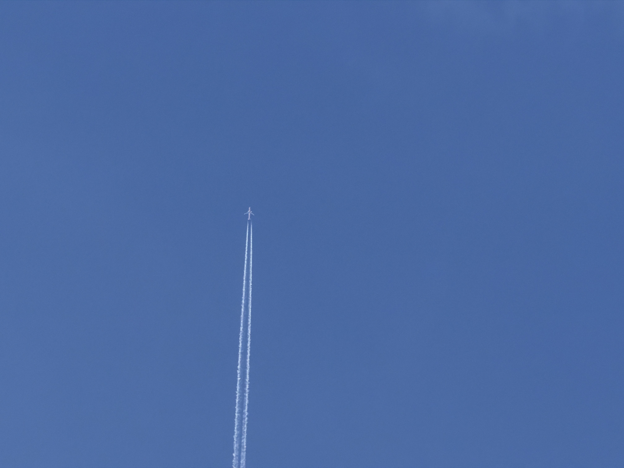 The image displays a clear, expansive blue sky above Poros, Greece, under bright daylight conditions. The sole subject is a small jet airplane, flying at a high altitude and positioned towards the upper-middle of the frame. The plane is depicted as a tiny white form against the deep blue, leaving a distinct, long white contrail that stretches vertically downwards from its position. Initially, the contrail appears as two parallel streams directly behind the aircraft, indicative of its engines, before slightly diffusing as it extends further. The sky is otherwise entirely cloudless, suggesting excellent weather. No other objects, people, or text are visible in this minimalist composition, which focuses on the distant aircraft and its atmospheric trace.