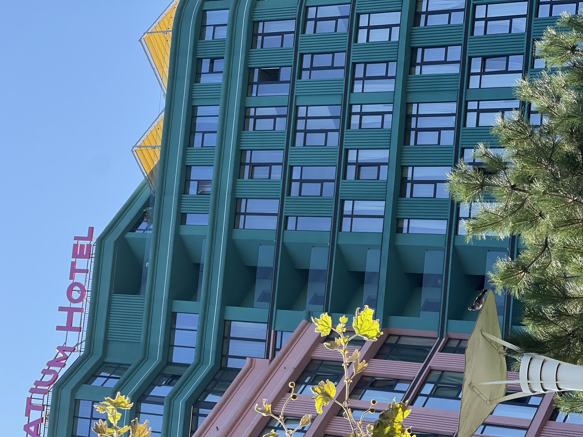 The image shows the exterior of the "ATIUM HOTEL" building in Odesa, Ukraine. The hotel features a distinctive teal-colored facade with a curved design, punctuated by numerous windows with dark frames. There are also balconies with glass railings on multiple levels. To the left of the teal building, a portion of a pink building is visible, also with windows. Above the teal building, two triangular yellow awnings are prominent against a clear blue sky. In the foreground, a lamp post with a white shade is partially visible, and a pigeon is perched on its surface. Green pine branches are also in the foreground, partially obscuring the lower parts of the buildings. The overall impression is of a modern architectural design in an urban setting.