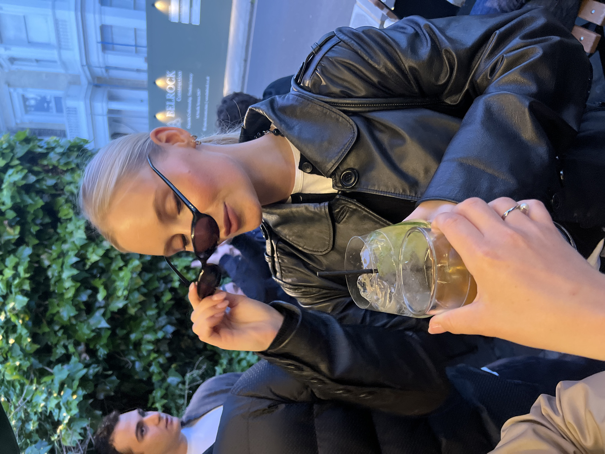 The image captures a young, fair-skinned woman with blonde hair, wearing a black leather jacket and sunglasses, and holding a glass of a cocktail with ice and a straw. She appears to be seated at an outdoor establishment. The edge of another person is seen on the right, holding the drink. The foreground includes foliage, possibly ivy, suggesting an outdoor seating area. In the blurred background, there is a building facade with a sign that reads "BELROCK Construction," indicating a construction company. Given the details, it is plausible that the location is in London, United Kingdom. The individuals appear to be casually socializing and enjoying drinks outdoors. A man with dark hair and a white shirt is partially visible in the lower left.