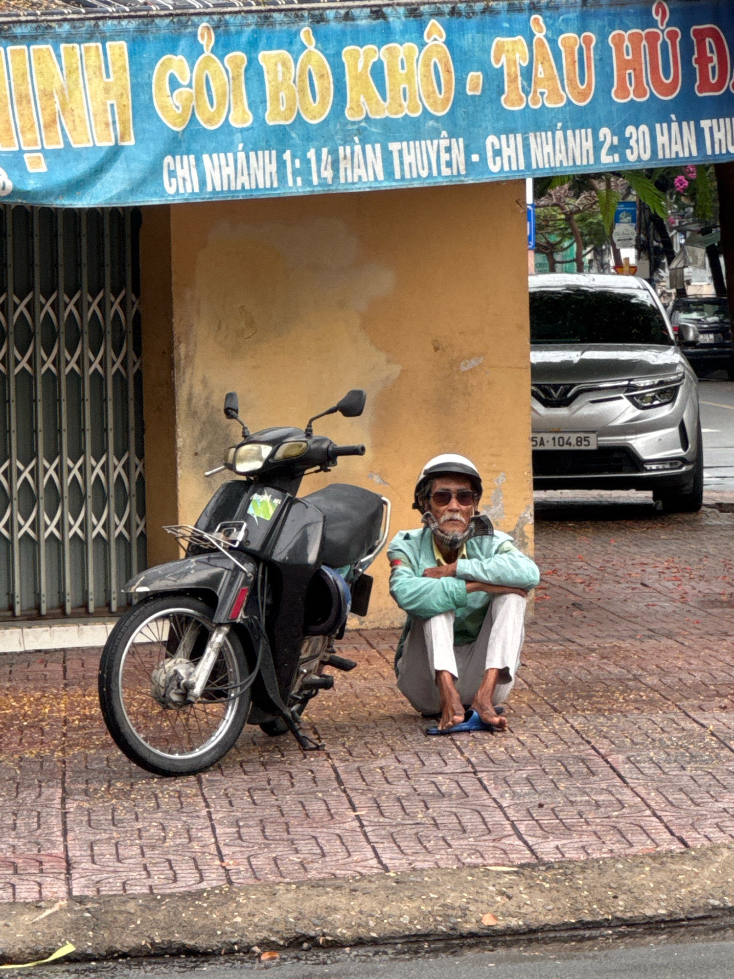An elderly man with a goatee, wearing a white helmet, sunglasses, a light green jacket, and white pants, sits cross-legged on a reddish-brown brick sidewalk in front of a yellow building. He is wearing blue flip-flops. A dark-colored motorcycle is parked to his left, with its front wheel visible. Behind the man and the motorcycle, a modern silver car with a Vietnamese license plate reading "5A-104.85" is partially visible. A blue banner with Vietnamese text and yellow lettering hangs above the scene, partially obscuring the yellow building. The banner reads "LINH GÔI BÒ KHÔ - TÀU HỦ ĐÁ" and "CHI NHÁNH 1: 14 HÀN THUYỀN - CHI NHÁNH 2: 30 HÀN THUYỀN". A closed metal gate with a criss-cross pattern is located to the left of the building. Fallen leaves are scattered on the sidewalk and the street. The scene appears to be in Nha Trang, Vietnam.